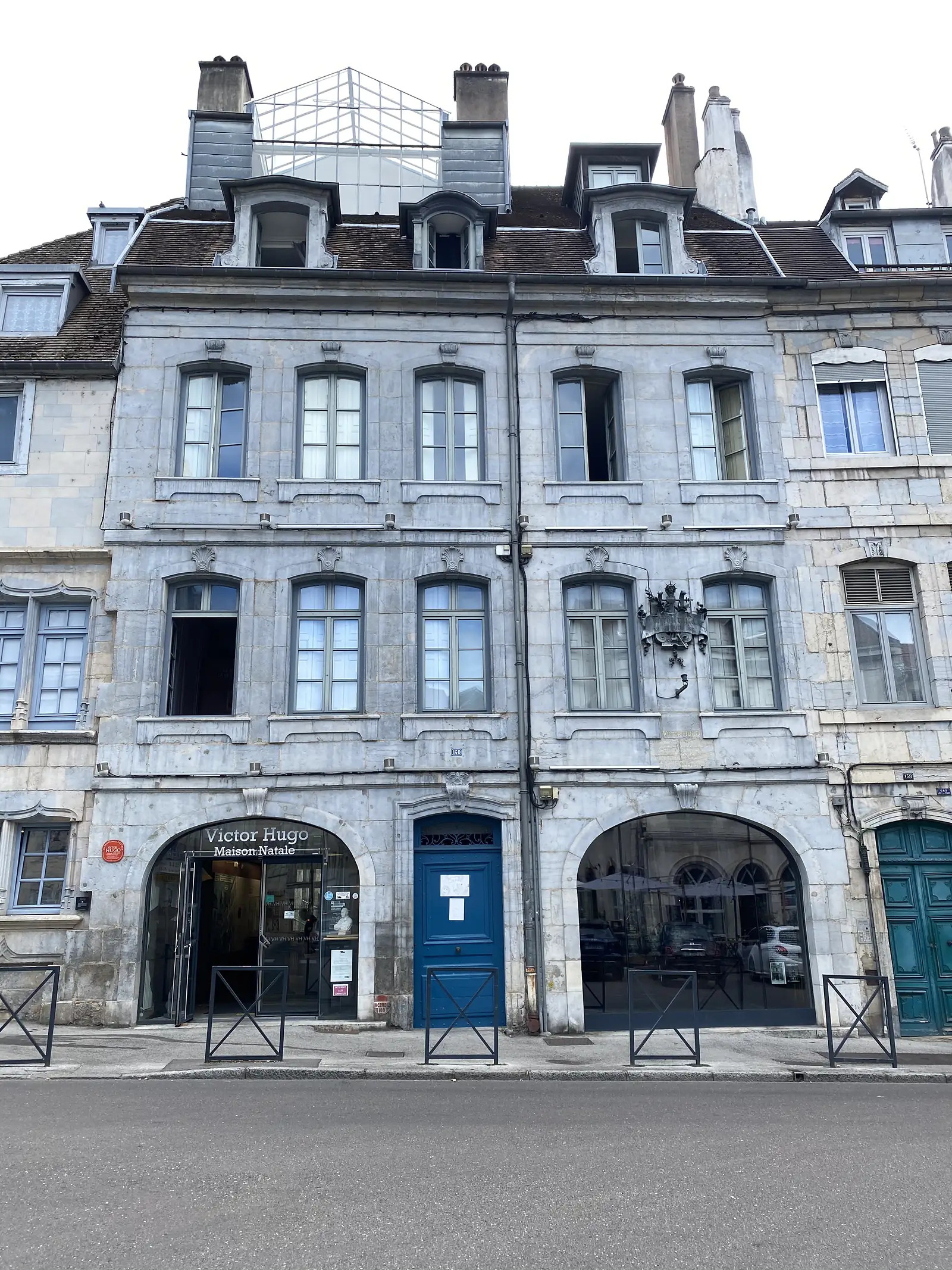 The stone facade of the Maison Natale de Victor Hugo in Besançon, its ground-floor arches now housing shops beneath dormered attic windows.