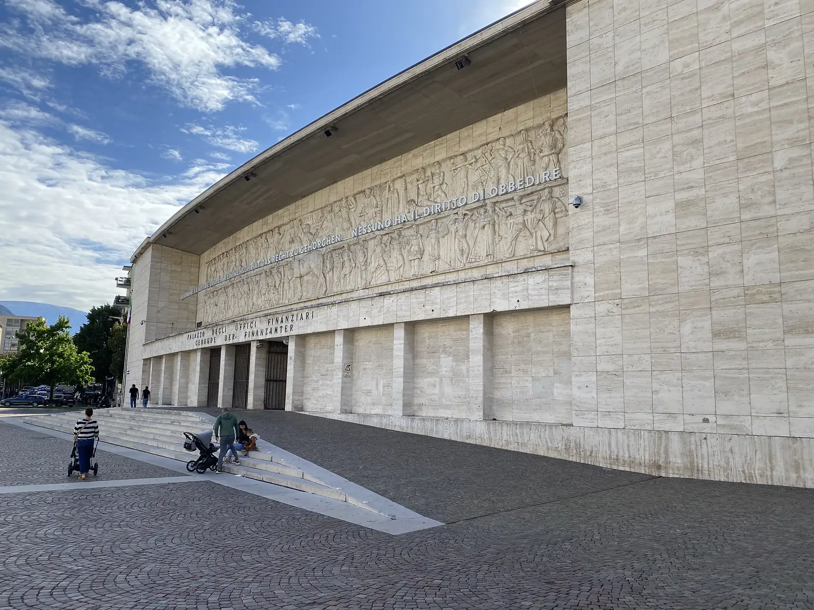 The curved travertine facade of a 1930s public building carries a huge carved frieze and the illuminated inscription "NESSUNO HA IL DIRITTO DI OBBEDIRE", above doorways lettered "PALAZZO DEGLI UFFICI FINANZIARI" and "GEBÄUDE DER FINANZÄMTER".