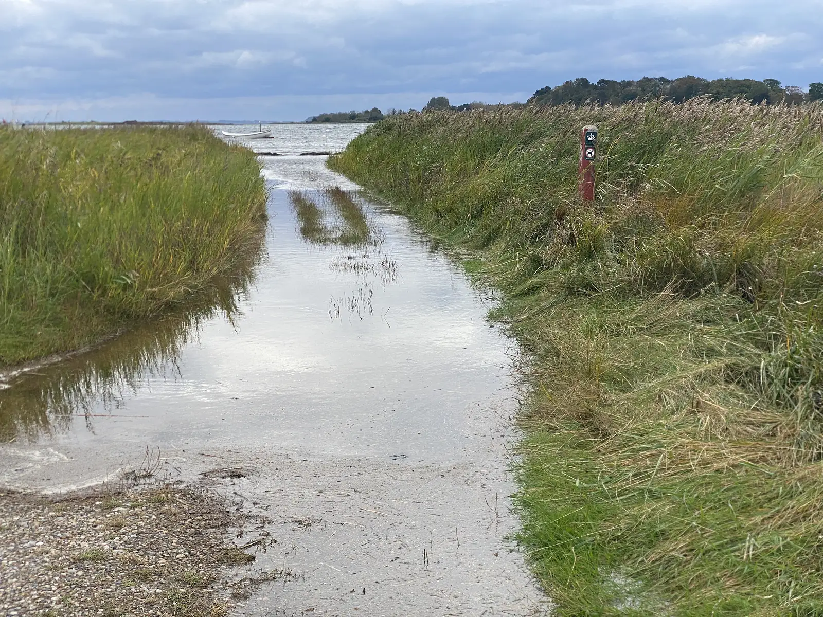 A sandy shore track flooded by the sea, running past reeds and a red waymark post to a small white boat moored offshore.
