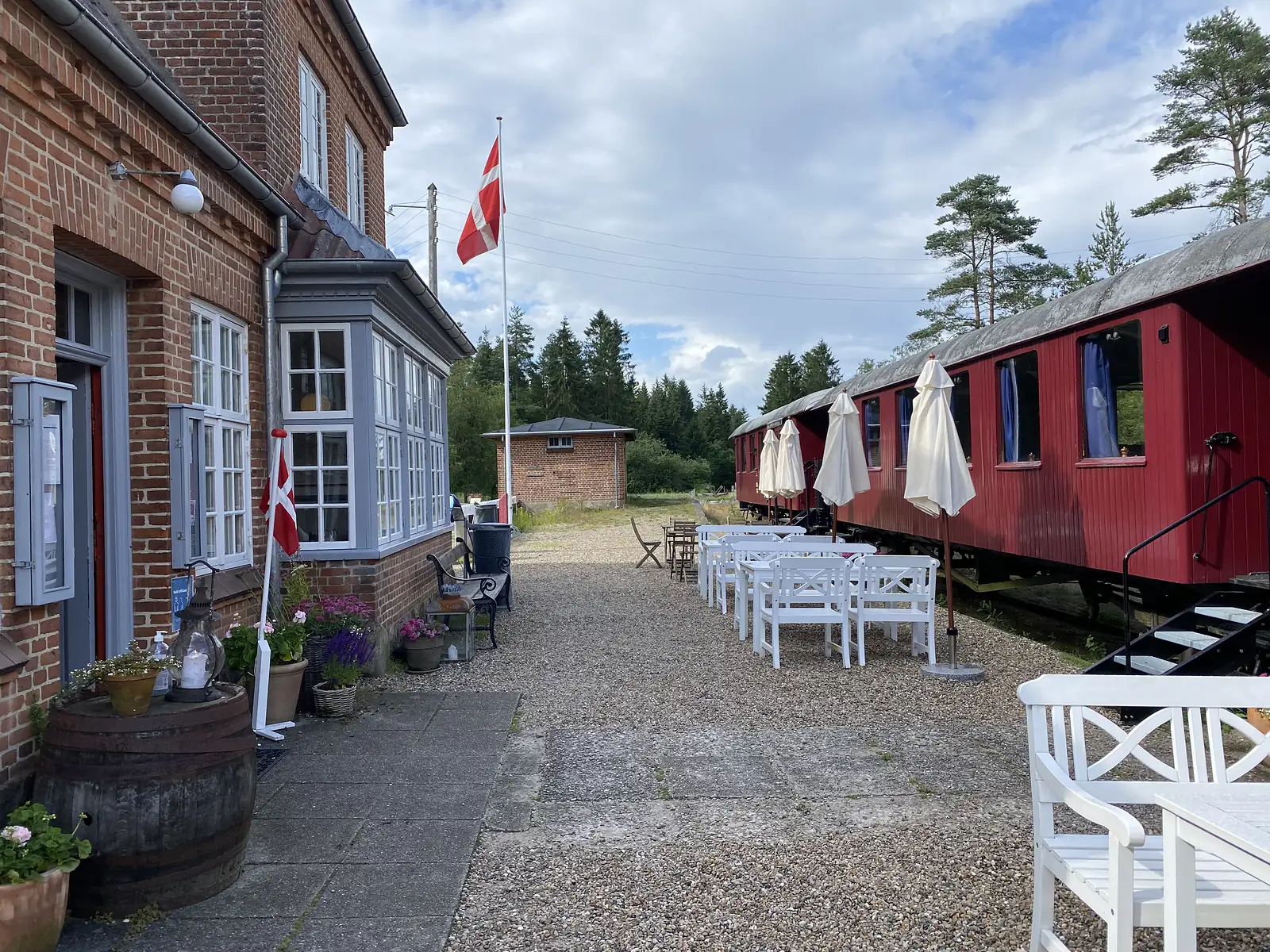 A red-brick station building with Danish flags and white cafe chairs on the gravel platform beside red wooden railway carriages.