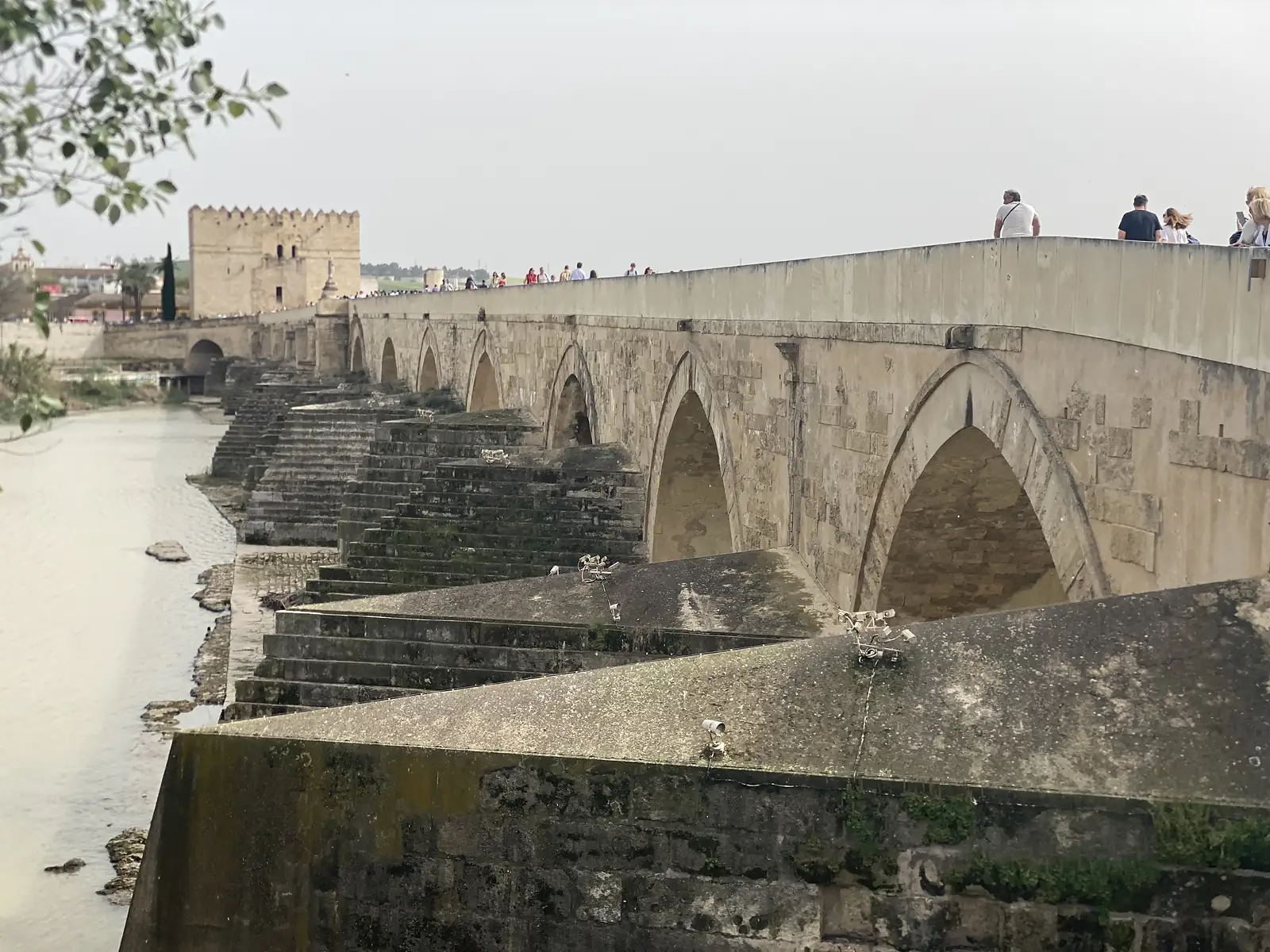 A long many-arched stone bridge crossing a wide river, people walking along it towards a battlemented gate tower at the far end.