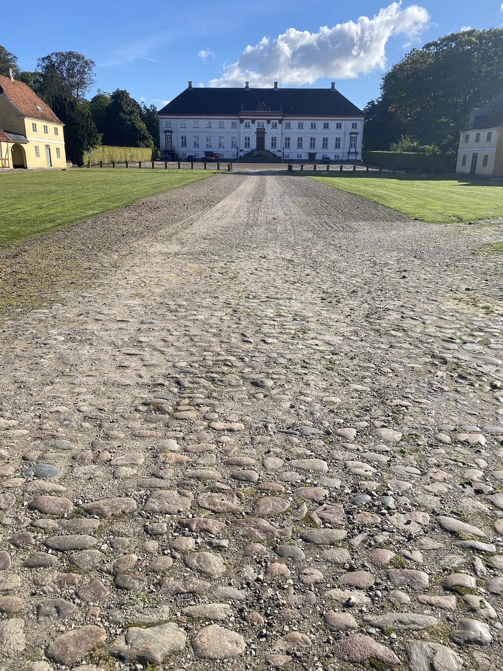 A cobbled avenue running dead straight across mown lawns to a white baroque mansion with a black mansard roof.