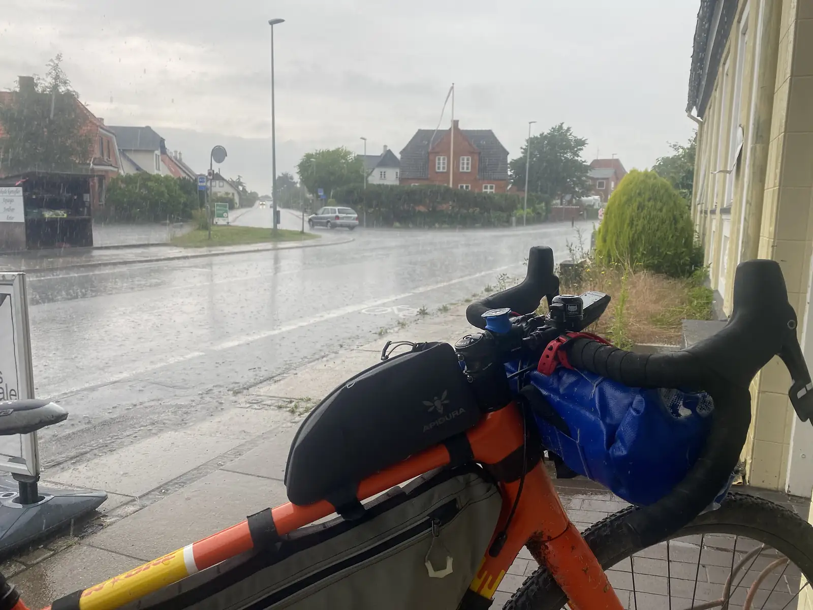 Sheltering from heavy rain under a shop awning, a loaded bike's handlebars in the foreground and rain bouncing off an empty road junction.