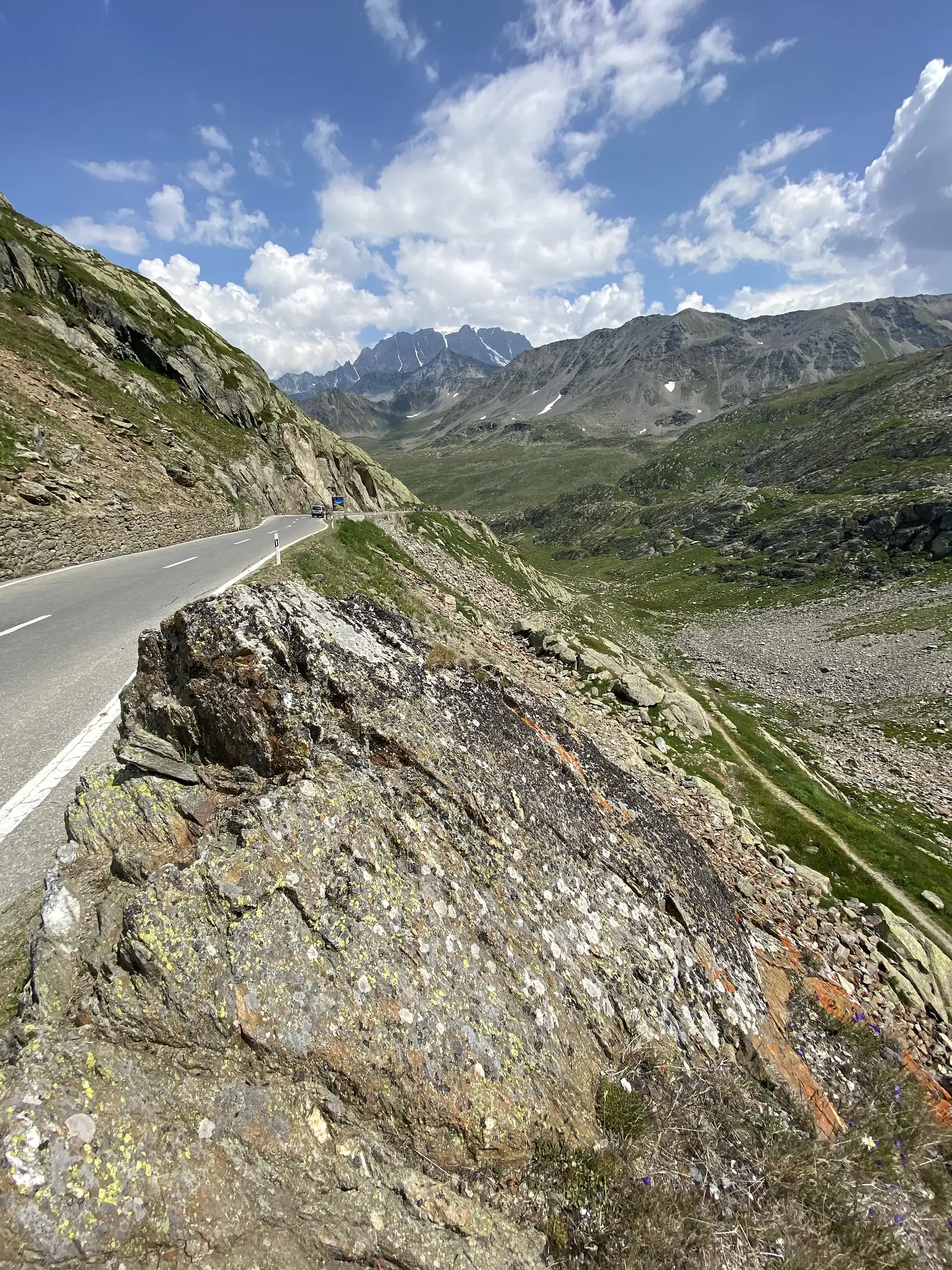 
A narrow mountain road hugging a lichen-covered rock face, with a wide scree-floored valley and distant peaks beyond.
