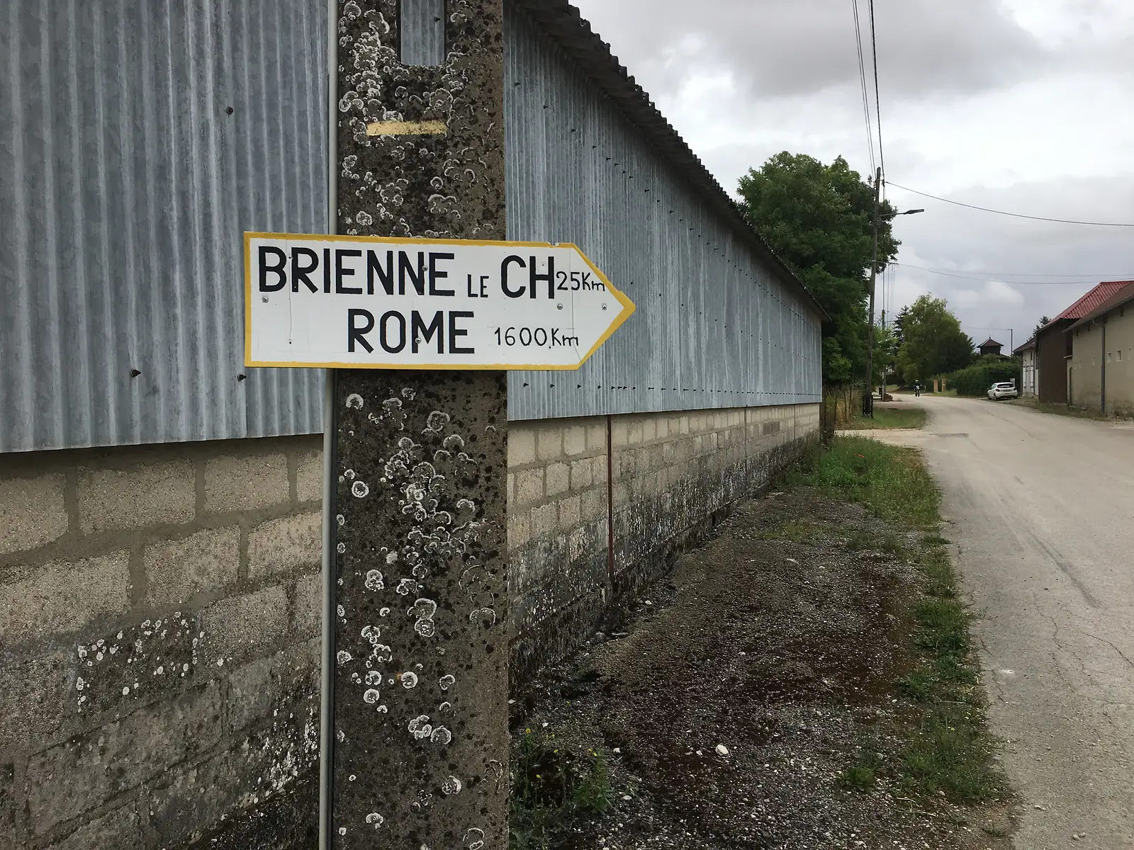 A hand-painted signpost reading BRIENNE LE CH 25km and ROME 1600km is fixed to a lichen-covered post against a corrugated-metal barn wall.