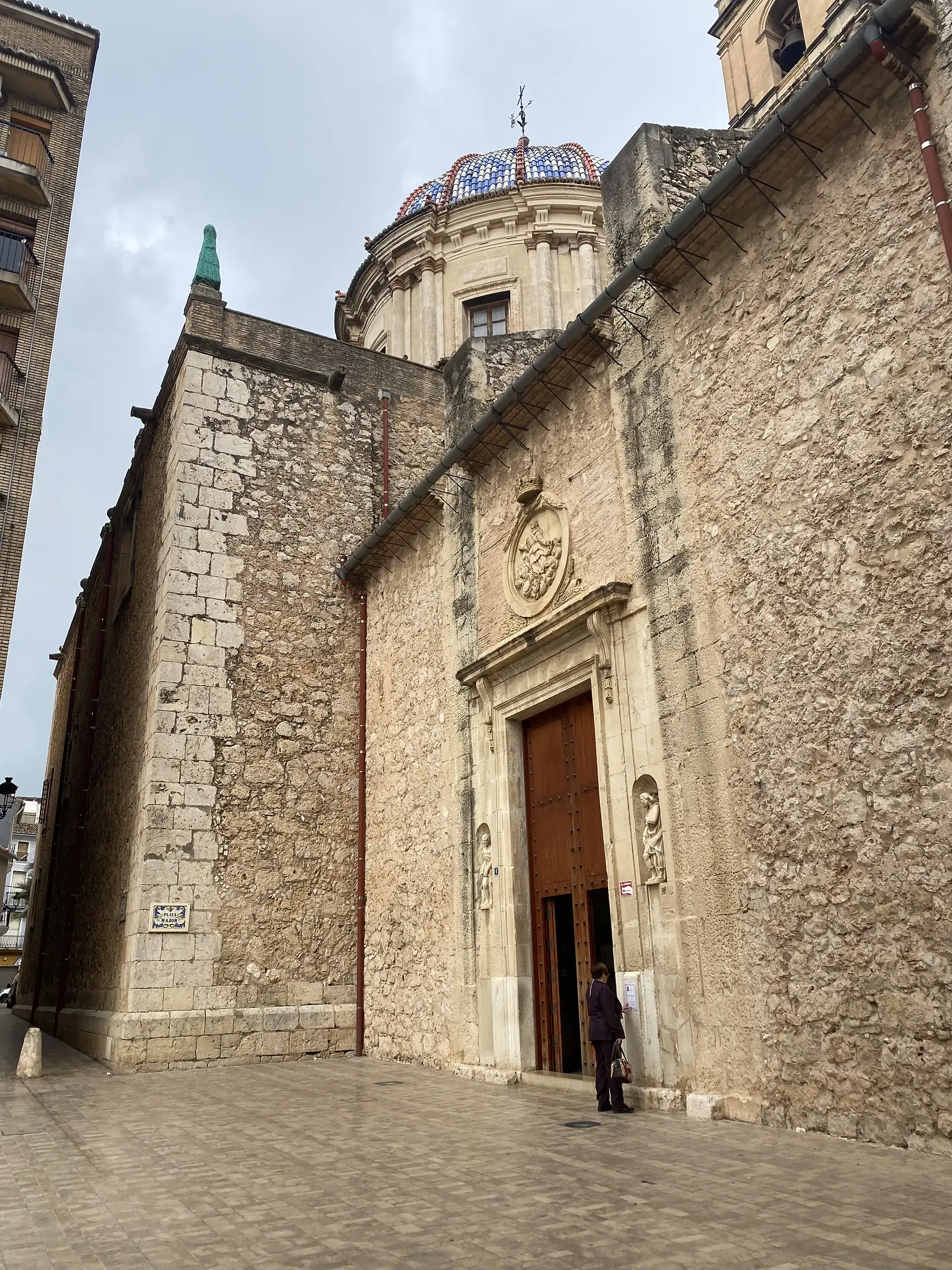 A massive rough-stone church front with a carved doorway and statues in niches, a blue-and-white tiled dome rising behind and a person at the open door.