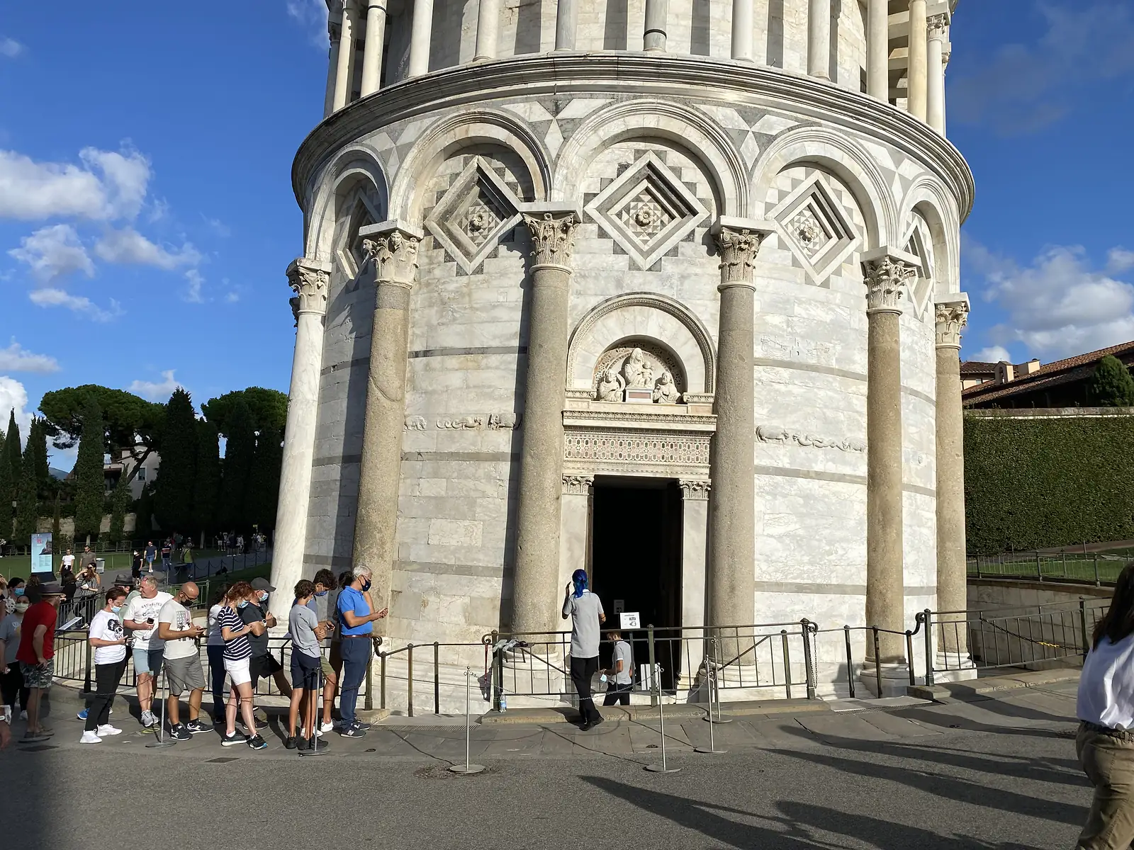 Visitors queue in face masks outside the round marble Baptistery of Pisa, waiting by its carved doorway beneath a relief of the Madonna and Child.