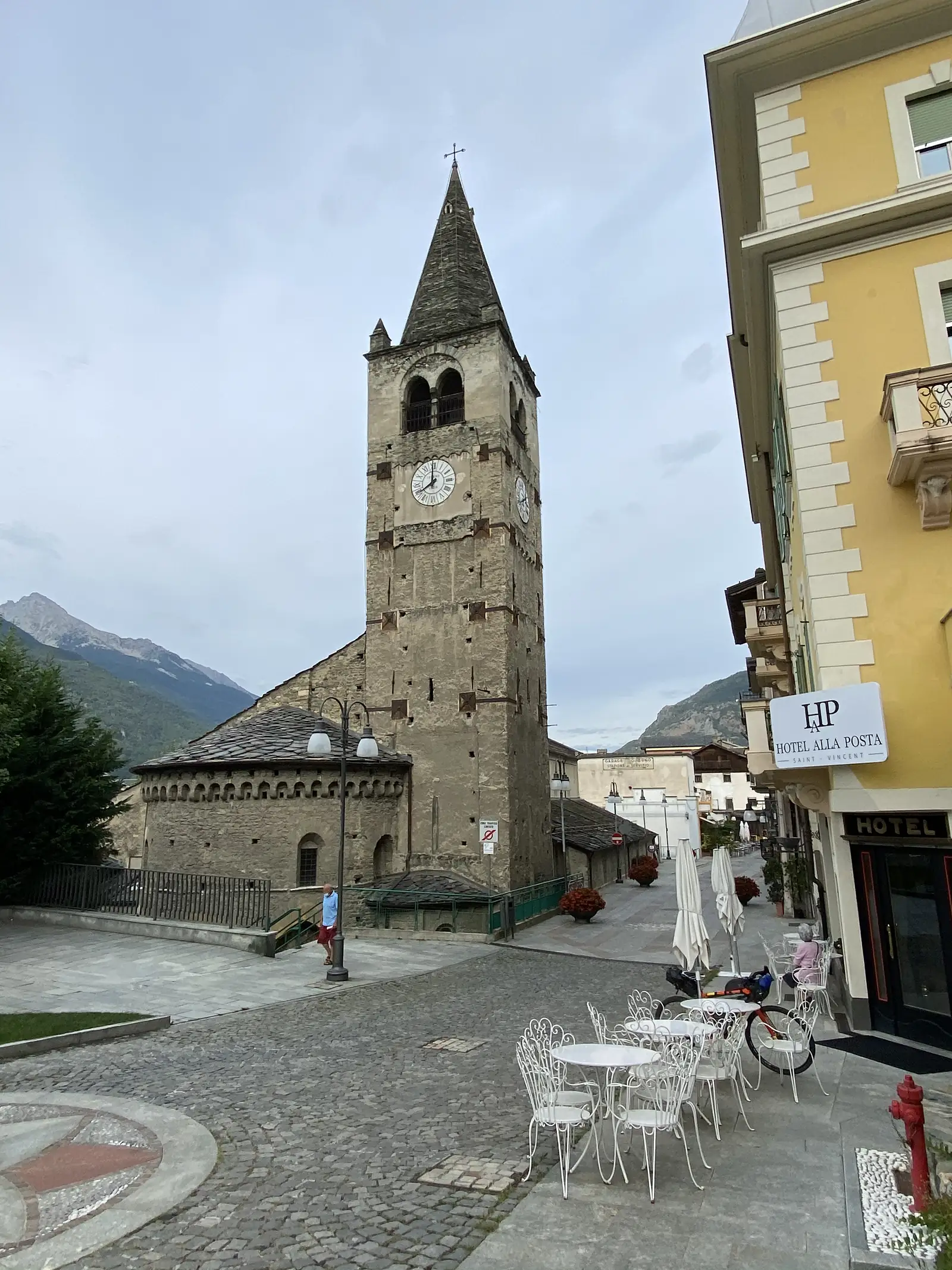 A stone Romanesque bell tower with twin clock faces rising beside a round apse, with mountains behind and cafe tables and a loaded touring bike parked at its foot.