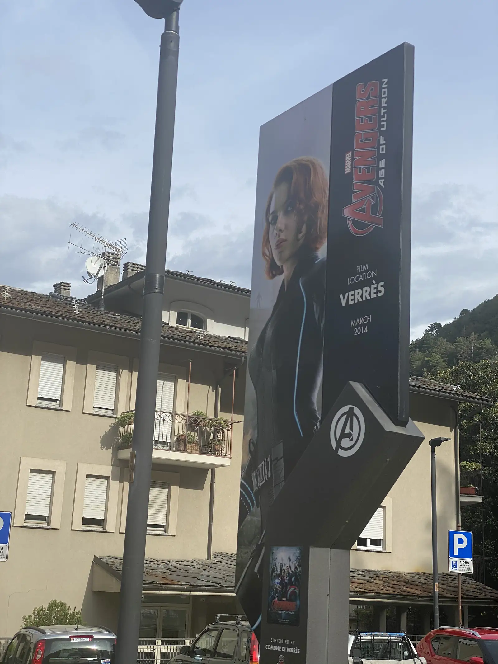 A tall street sign in Verrès showing a film-location poster for Marvel's Avengers: Age of Ultron, reading "Film Location Verrès, March 2014", against a backdrop of pastel apartment buildings.