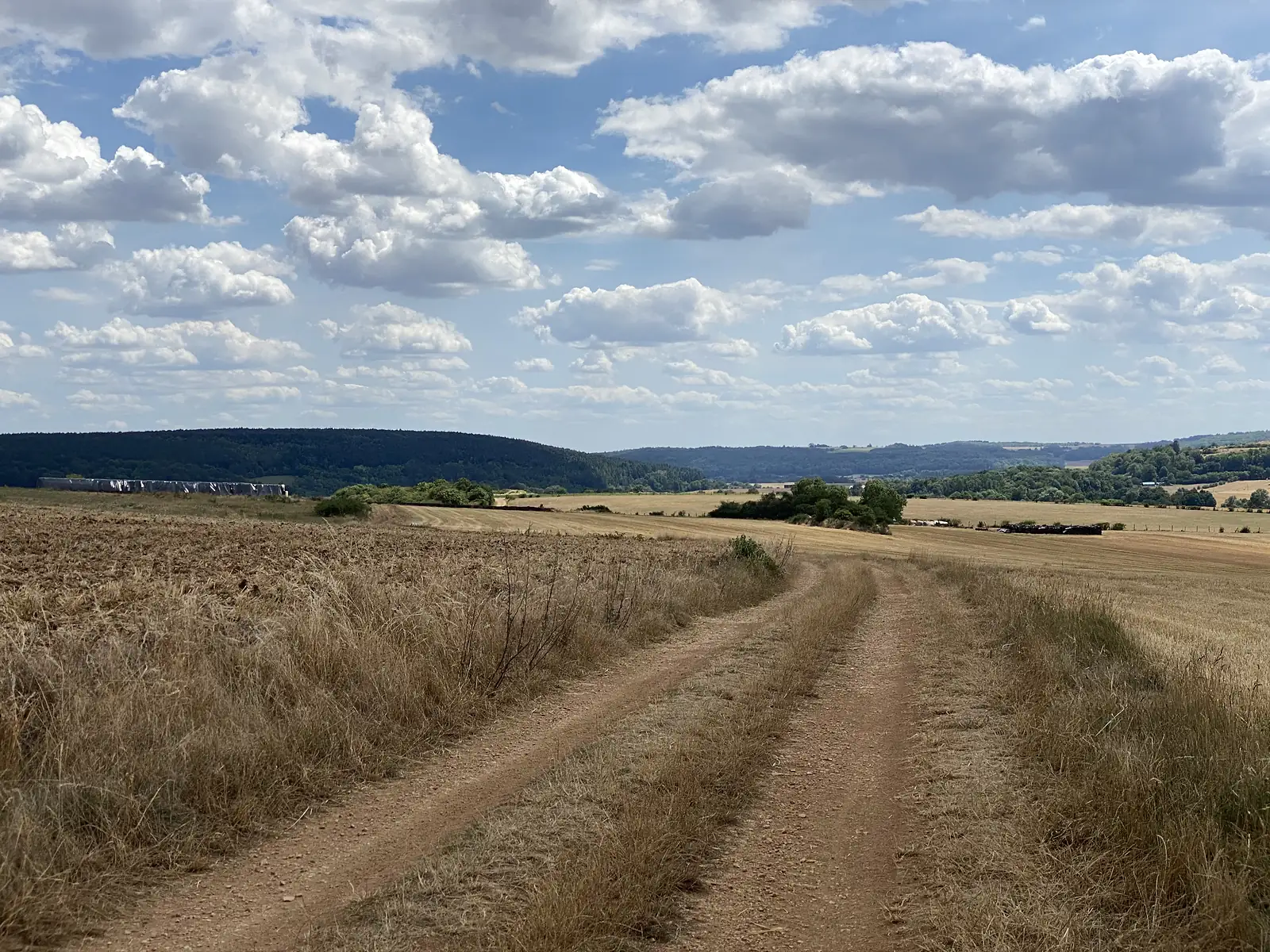 A twin-rutted dirt track crosses open stubble fields toward wooded hills, with hay bales and farm sheds scattered across the landscape below a storm-lit sky.