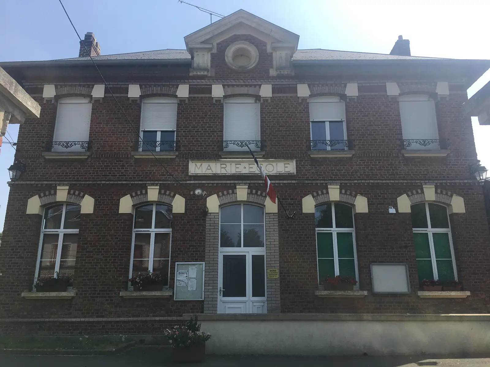 A red-brick village mairie-école building with shuttered upper windows and a tricolour flag hanging beneath its Mairie Ecole nameplate.
