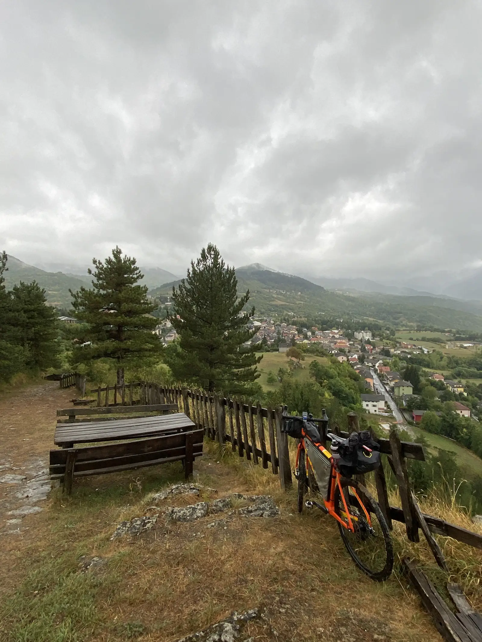 A laden orange gravel bike rests against a picnic-area fence beside pine trees, with a red-roofed village spread across the valley below.