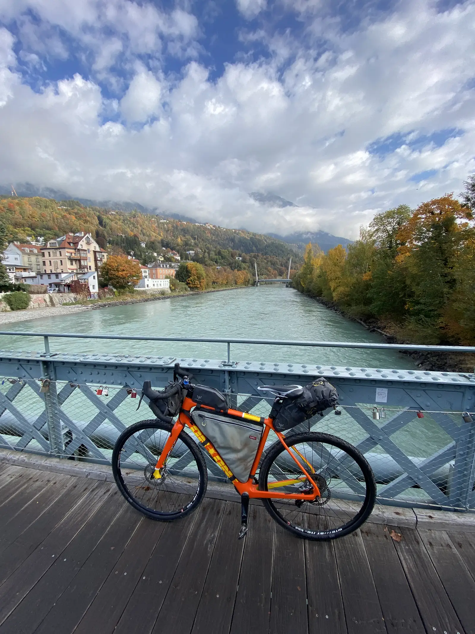 A loaded orange gravel bike stands on a steel truss footbridge over a pale green river, with autumn hillsides and riverside houses beyond.