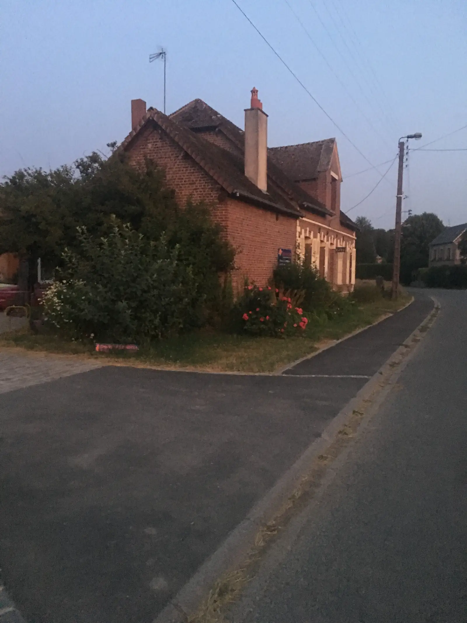 A brick house with ornate chimneys and a small spire stands beside a walled cemetery of pale headstones and a flowering rose bush, photographed in the soft light of early morning.