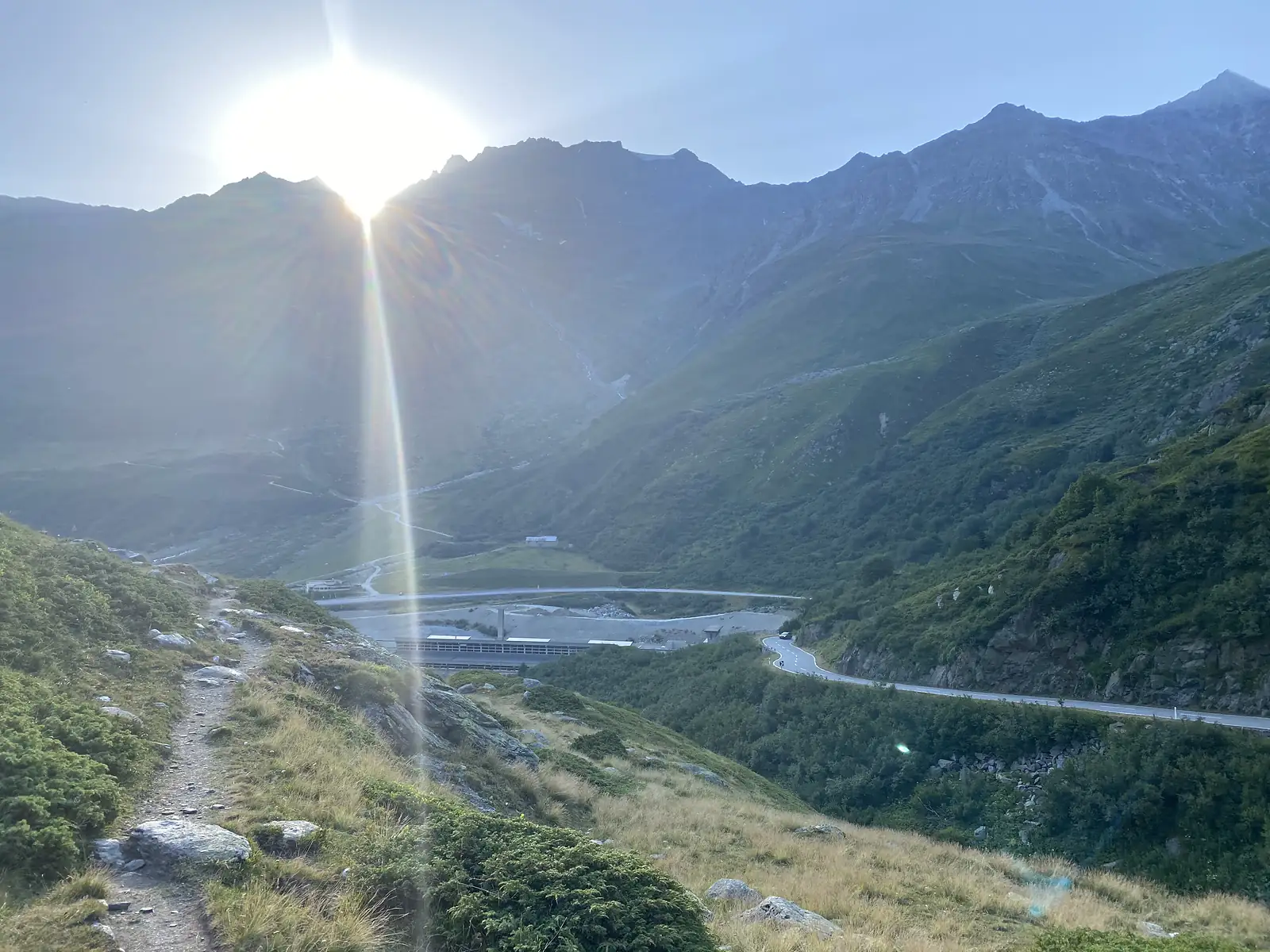 The sun breaks over a mountain ridge above a stony path descending towards a road and a cluster of buildings in the valley below.