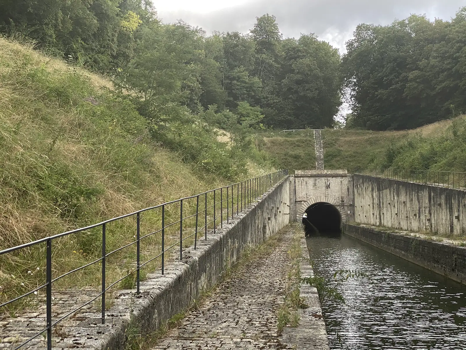 A canal towpath runs beside a stone-arched tunnel entrance set into a wooded hillside, railings following the water on both sides.