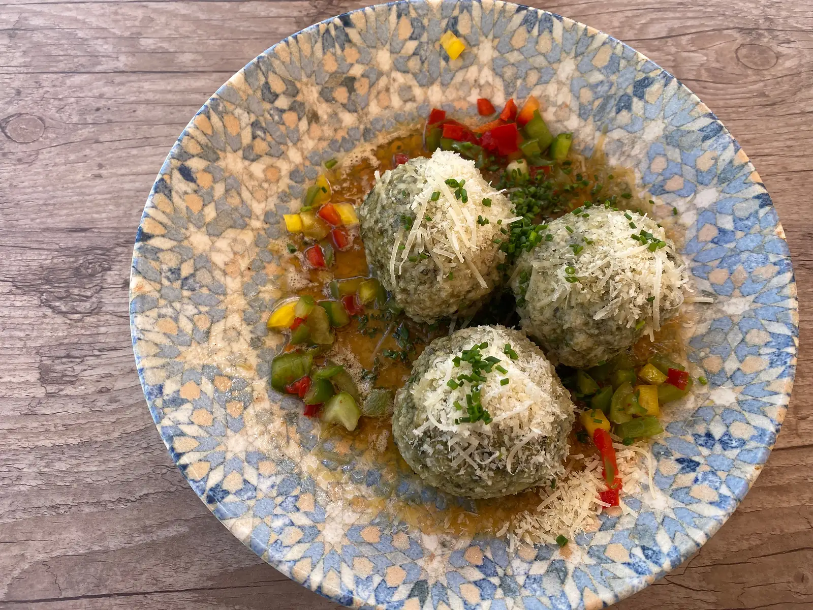 Three spinach dumplings dusted with grated cheese and chives sit in buttery sauce with diced peppers, in a blue-patterned bowl on a wooden table.