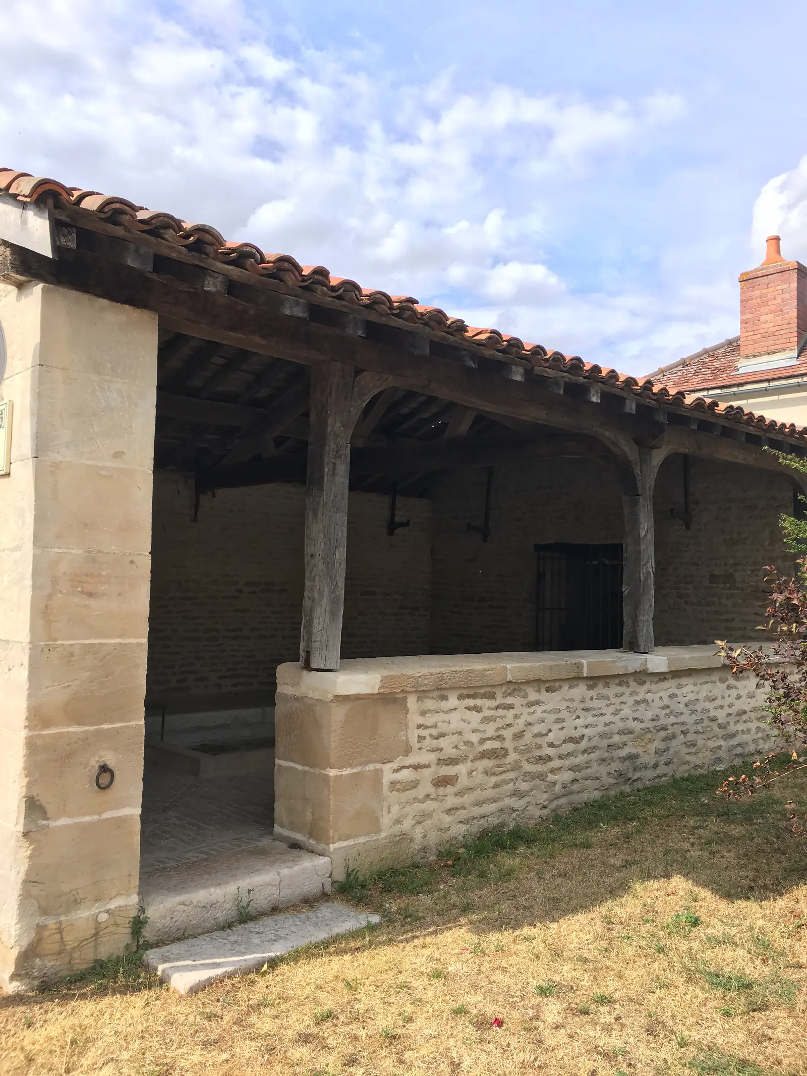 Covered stalls with a tiled roof and heavy timber posts shelter a stone trough at a village wash house.