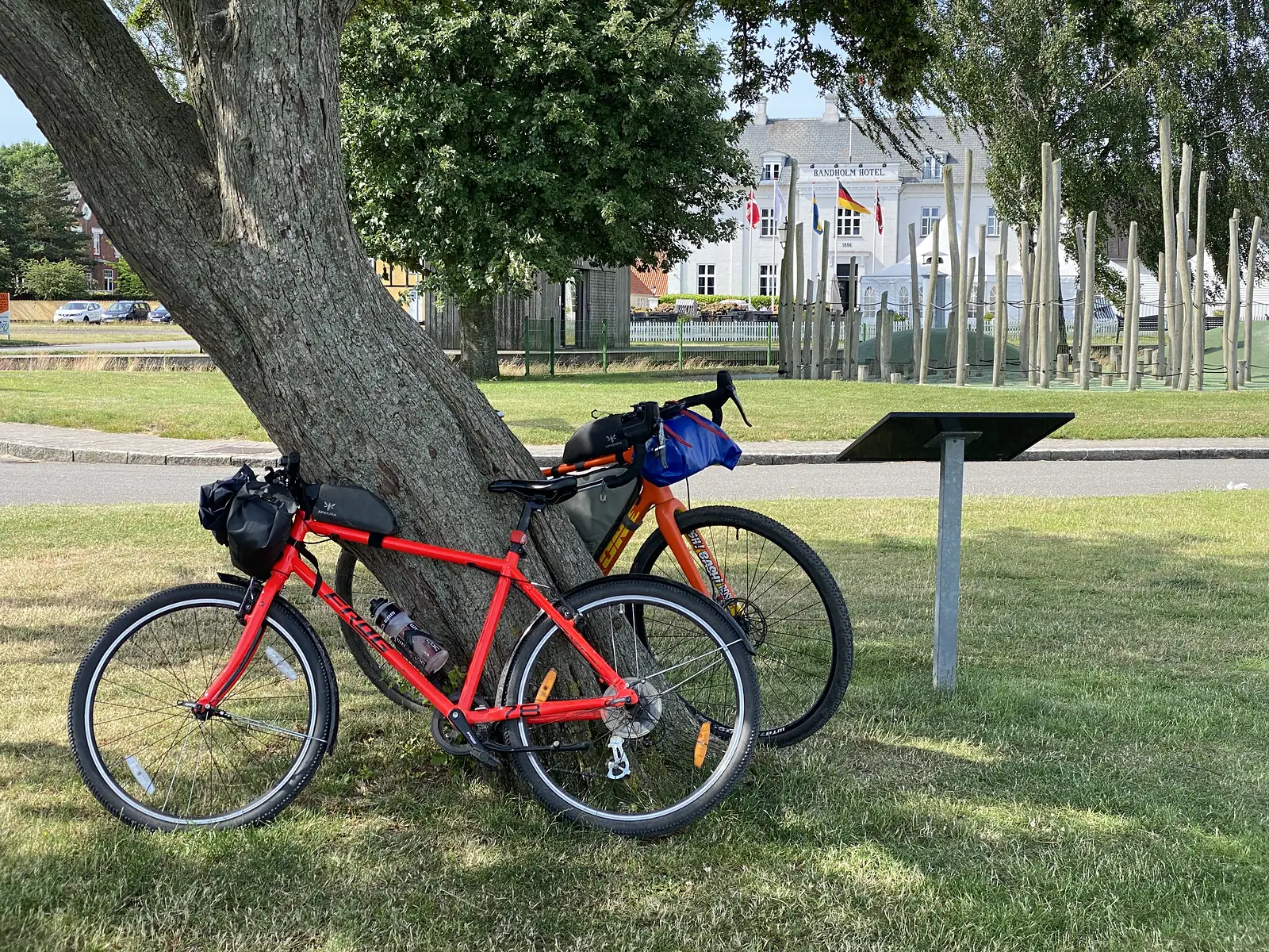 Two loaded bikes propped against a leaning tree on parched grass, with a white hotel lettered "BANDHOLM HOTEL" flying flags behind.