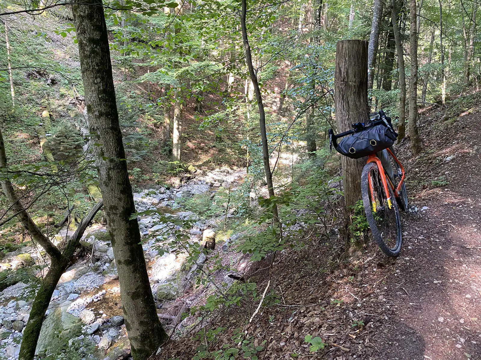 A laden orange gravel bike propped against a wooden post on a forest trail above a rocky, tree-lined stream.