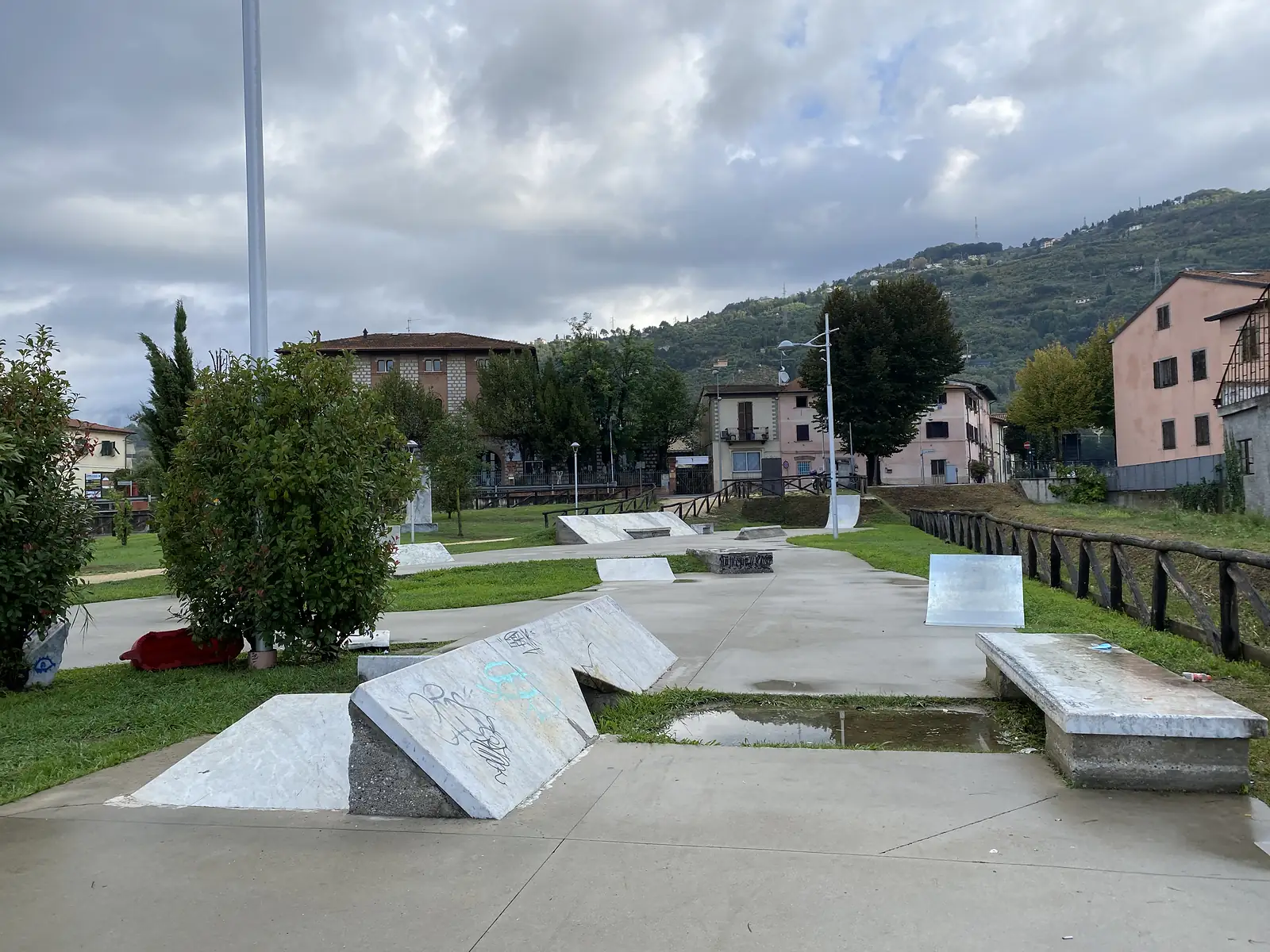 A graffiti-tagged concrete skatepark with marble-topped ramps and benches sits beside a stream, overlooked by pink and terracotta apartment buildings and wooded hills.
