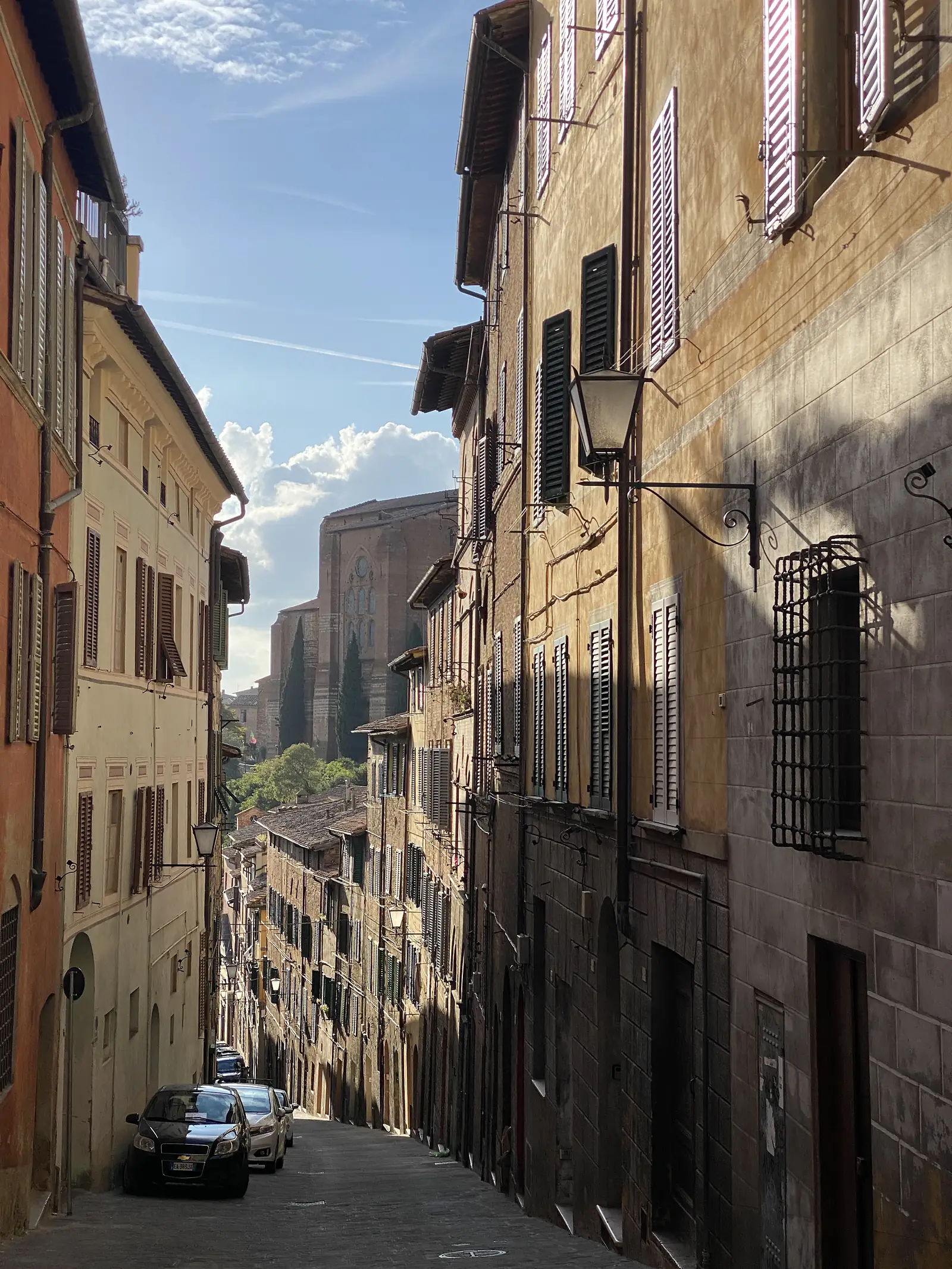 A narrow street lined with tall shuttered townhouses and wrought-iron lamp brackets, leading towards a distant tower, with cars parked along the kerb.
