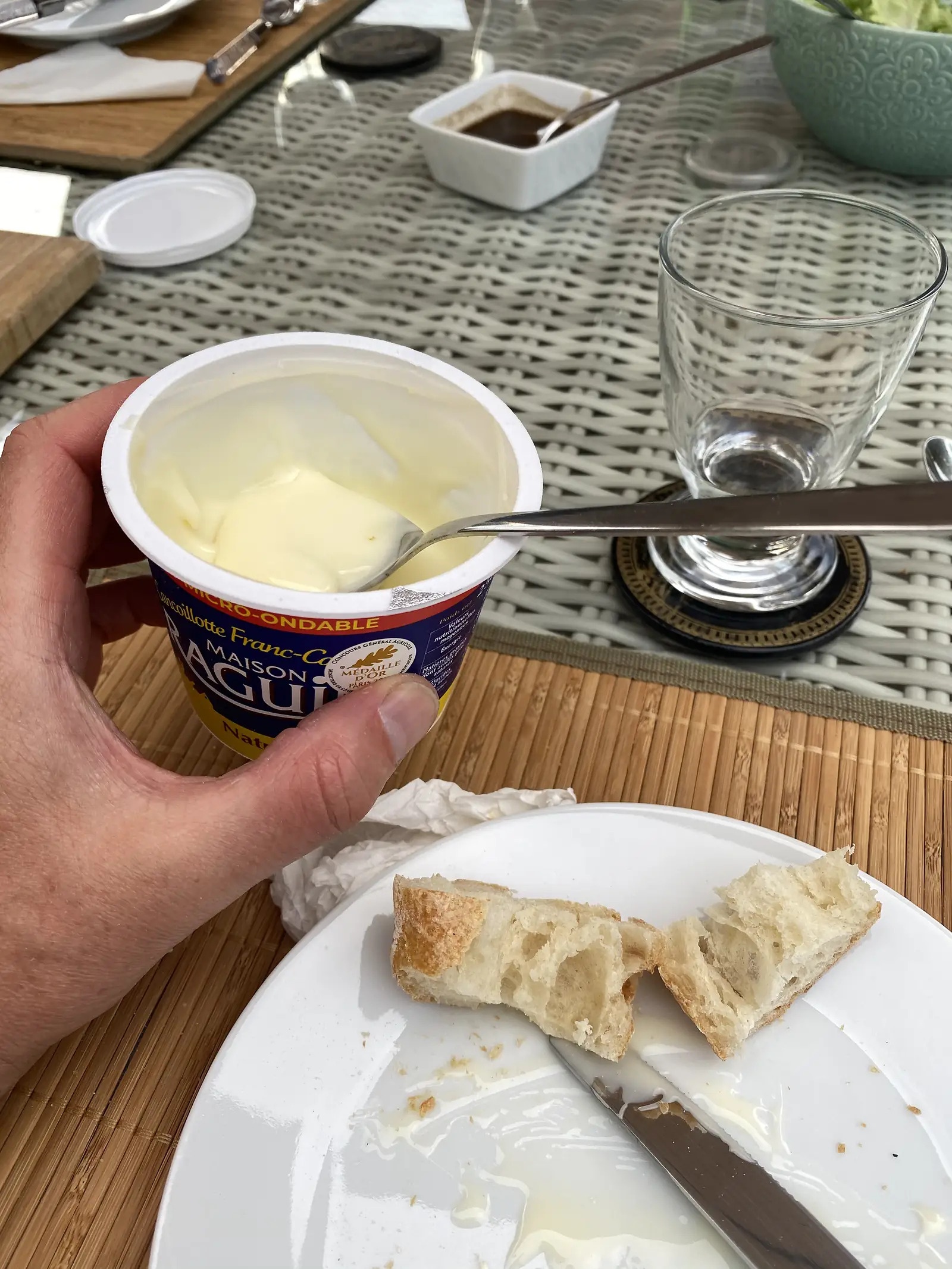 A hand holds an open tub of cancoillotte cheese with a spoon, ready to spread it on torn bread pieces set on a plate at an outdoor table.