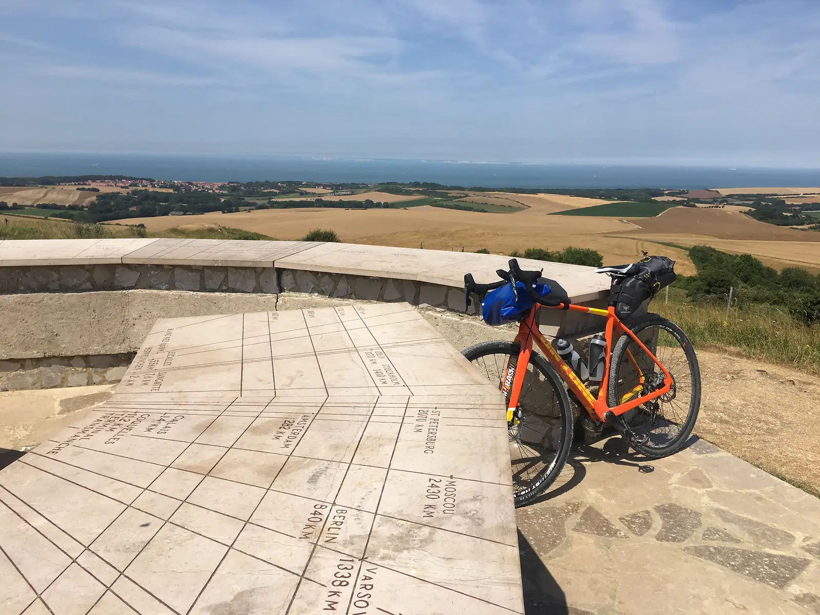 An orange bike leaning against a stone viewpoint wall inscribed with an orientation table listing distances to cities such as Calais, Berlin and Moscow, overlooking patchwork fields and the sea.