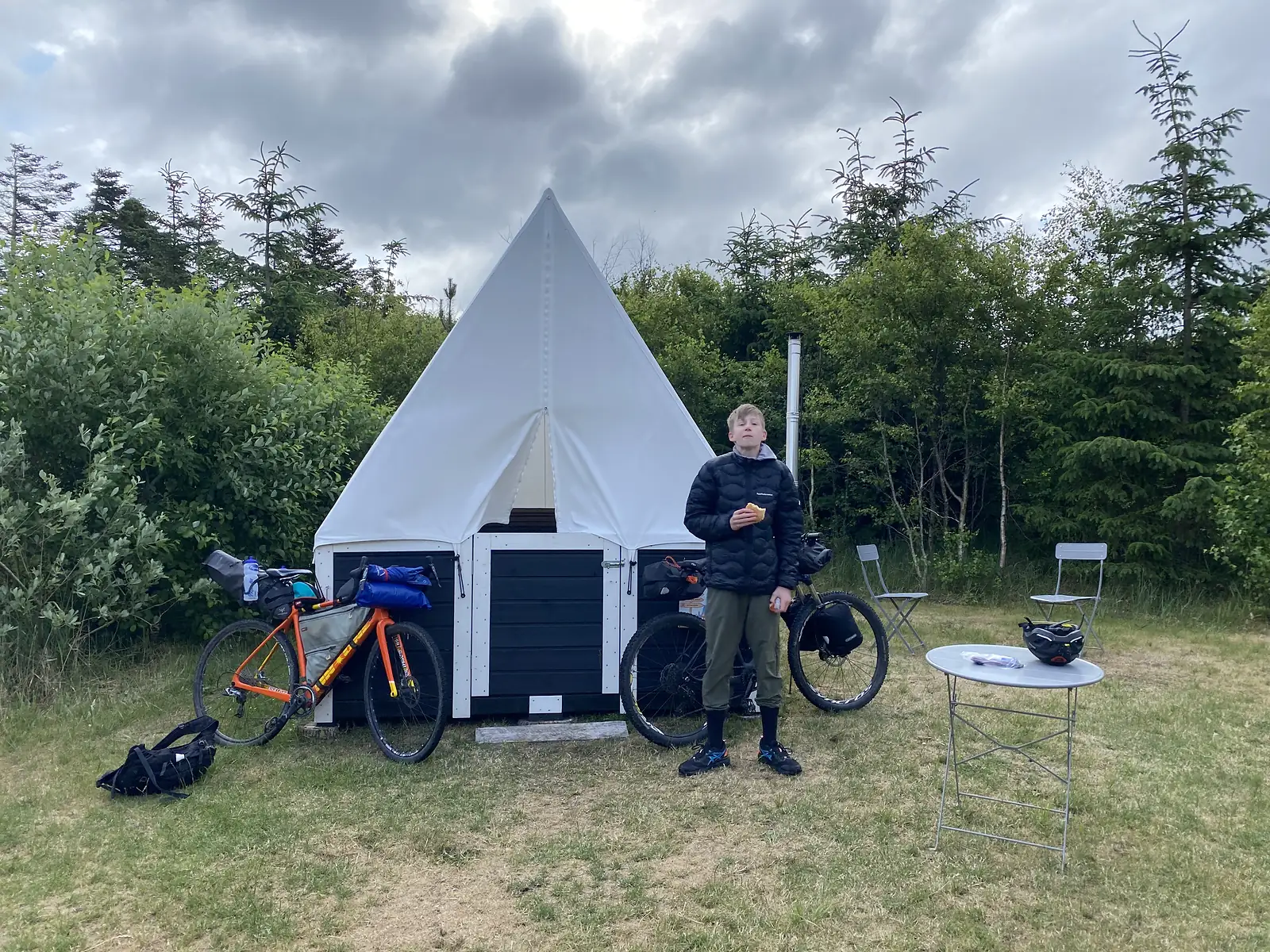 A young person in a black puffer jacket eating a pastry beside a loaded orange gravel bike outside a white canvas-topped camping shelter.