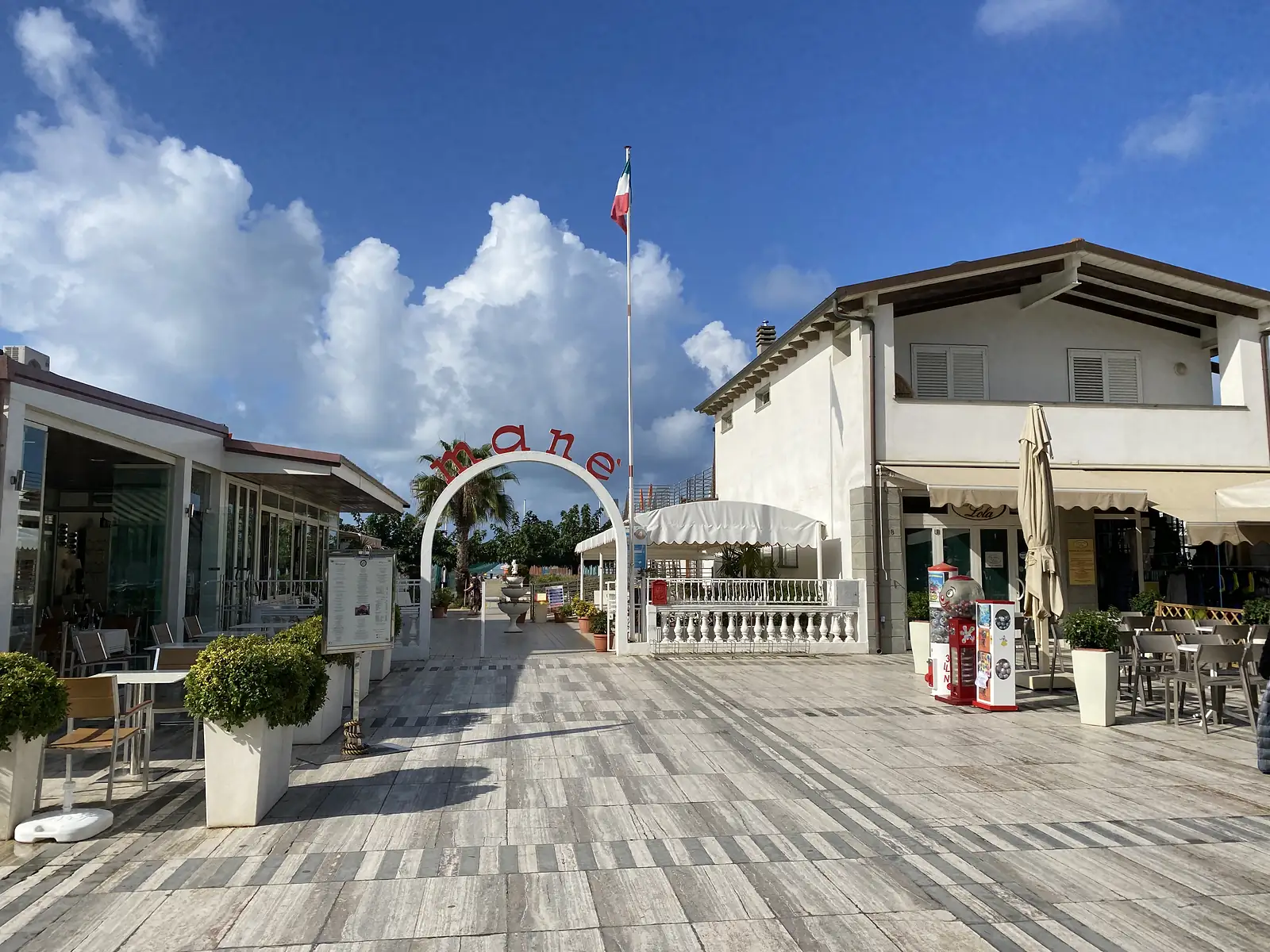 An archway entrance with lettering reading 'mané' leads into a beachfront courtyard flying the Italian flag, flanked by a café terrace and gumball machines.