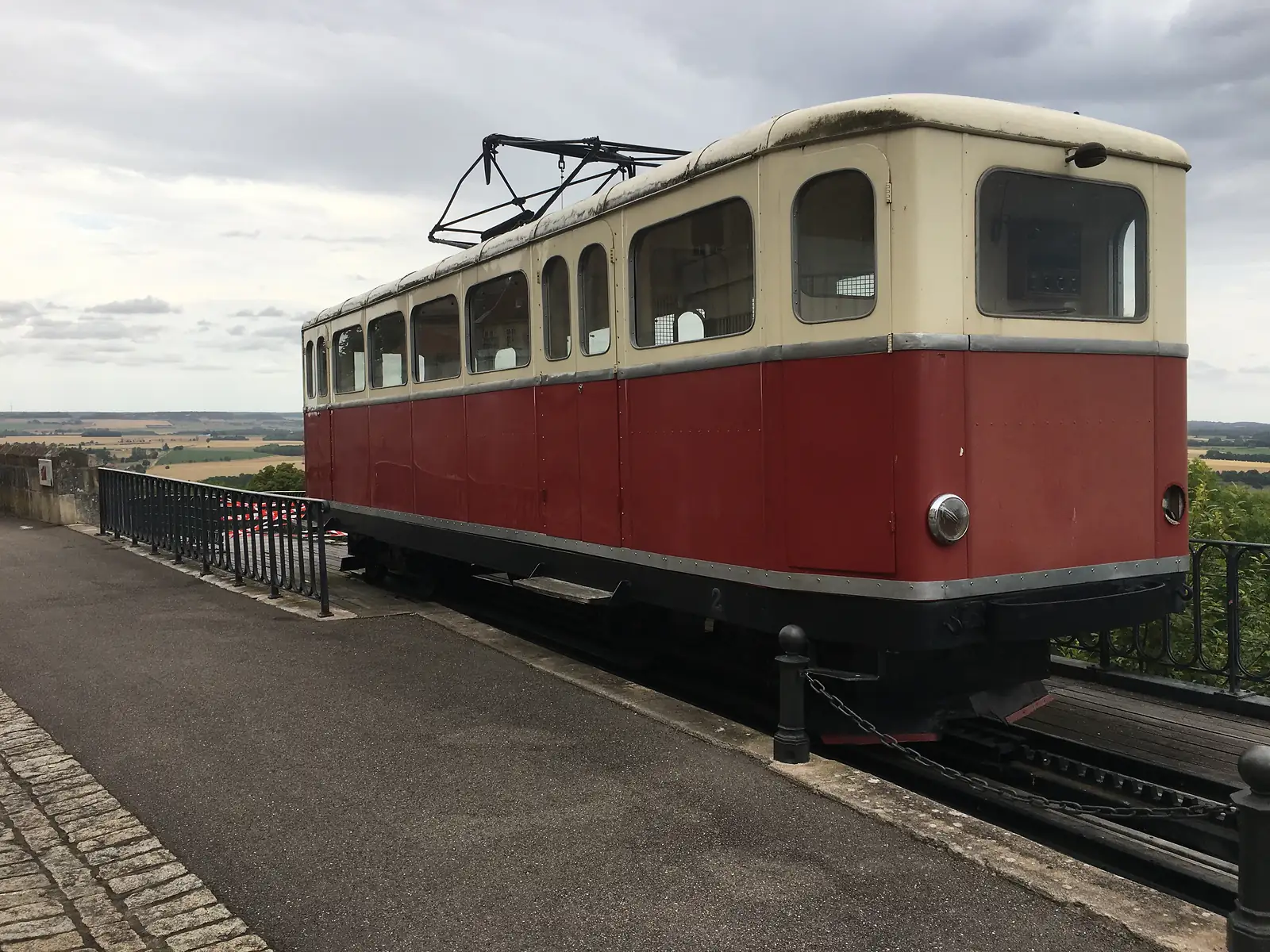 A vintage red-and-cream funicular tram car standing at a hilltop platform overlooking the Langres countryside, its trolley pole raised to overhead wires.