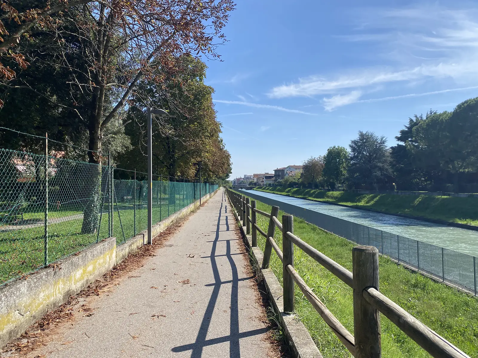 A tree-lined tarmac path runs beside a green canal behind a wooden fence, with houses on the far bank.