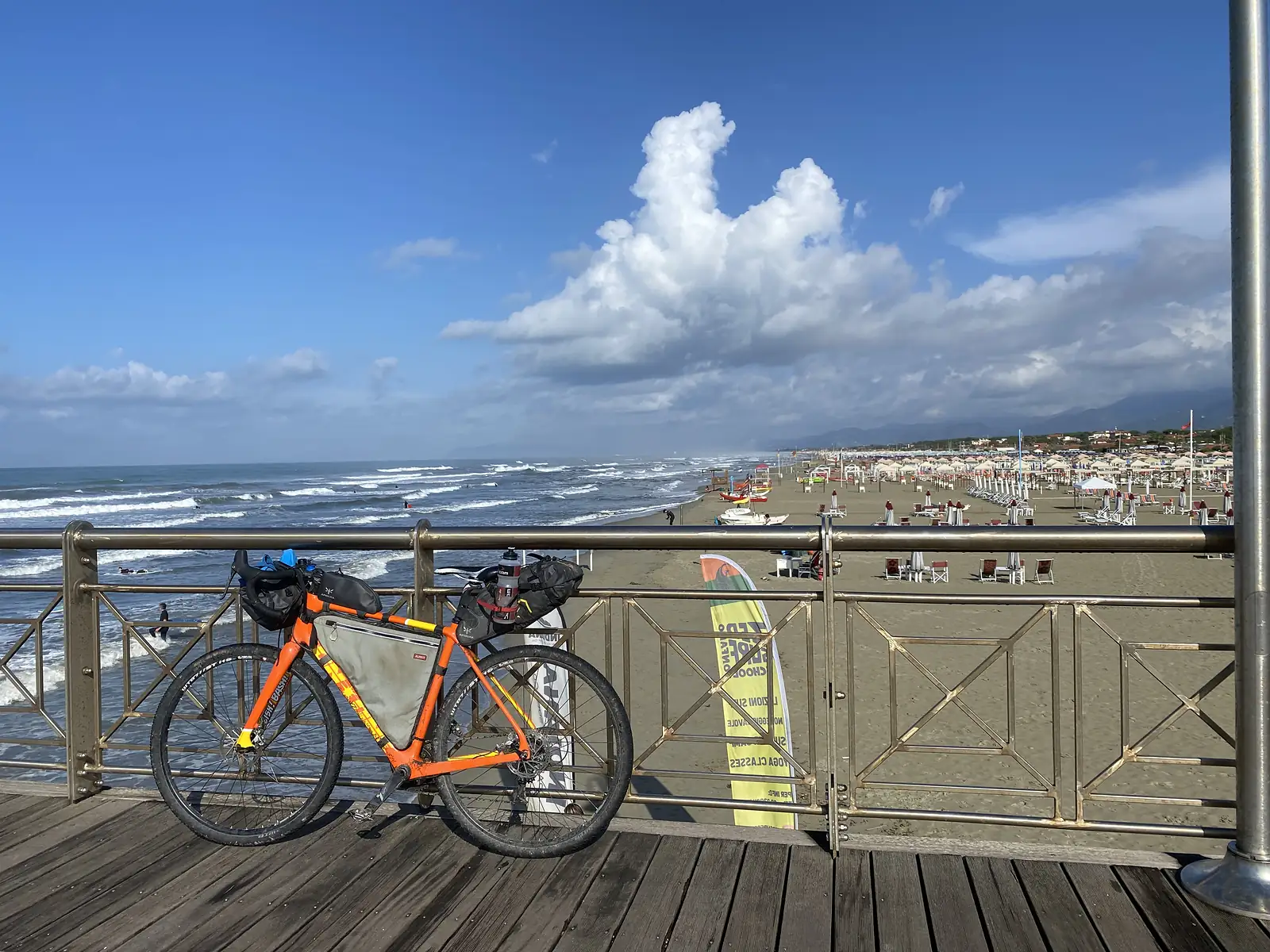 A laden orange gravel bike rests against a seafront railing, its frame bag and handlebar roll visible, with surfers in the water and umbrella-covered sand beyond.