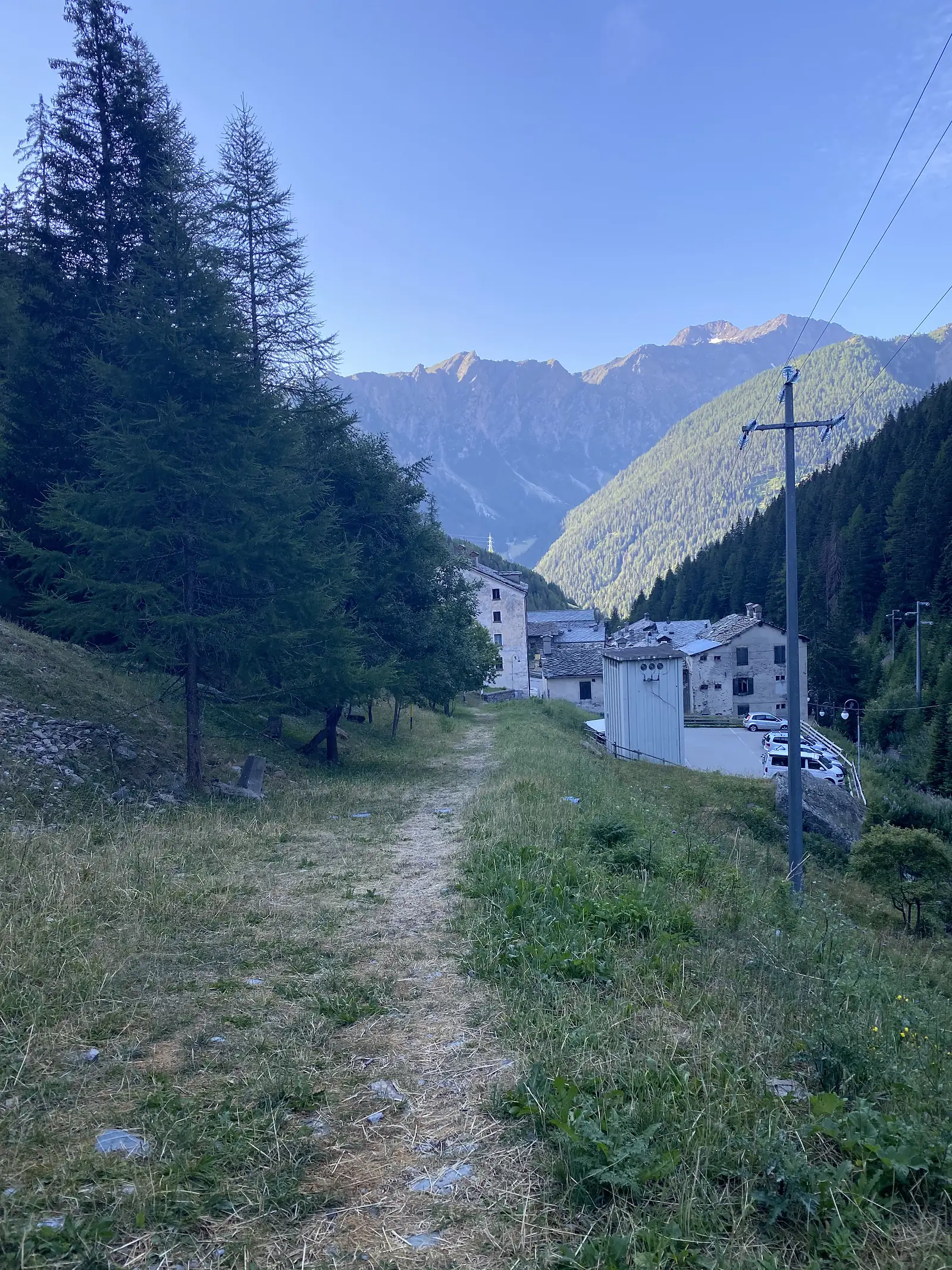 A grassy footpath leads down through pine trees toward a cluster of stone-built alpine houses, with parked cars and forested mountains rising behind.