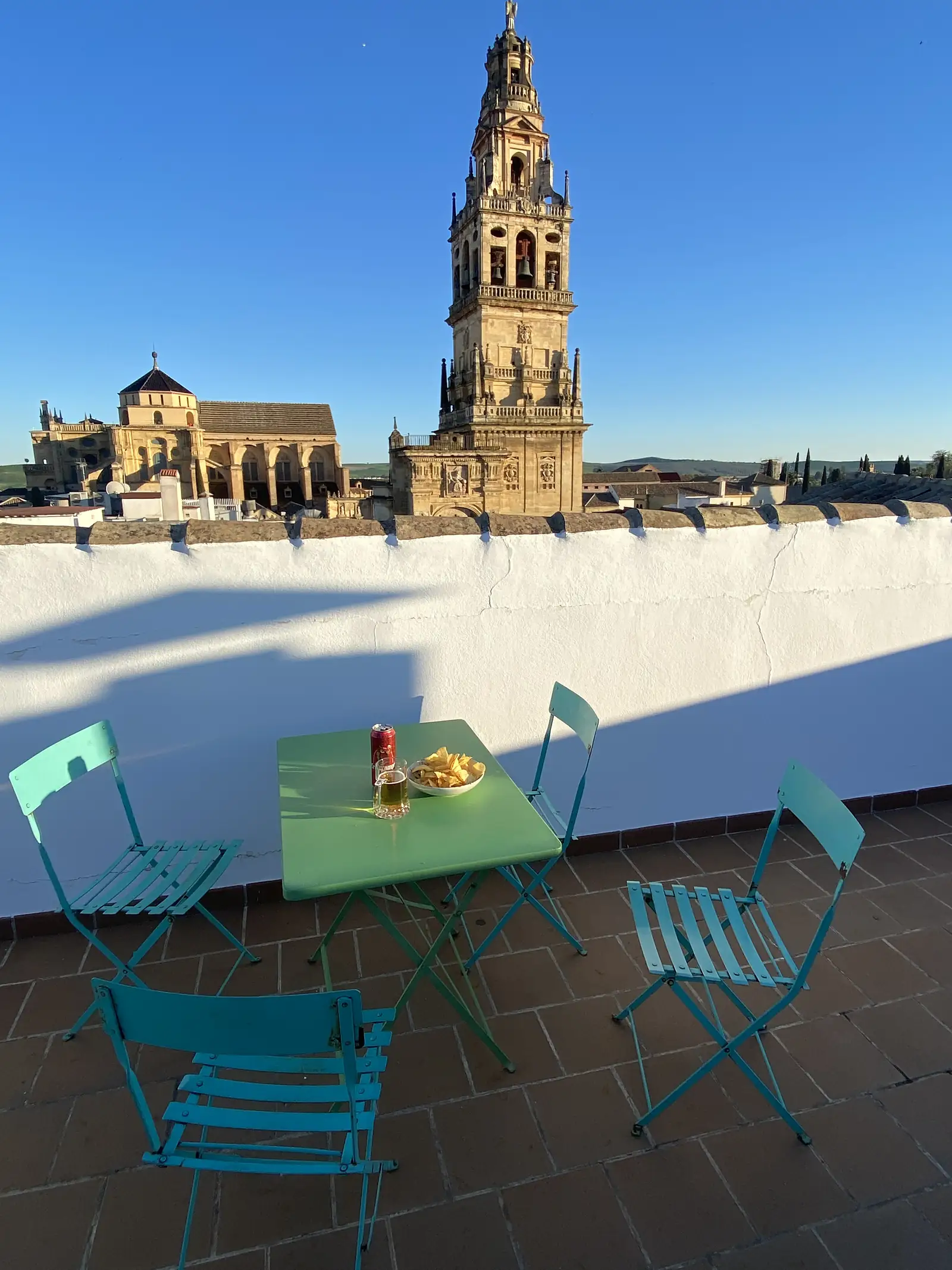 A beer and a bowl of crisps on a green table with turquoise folding chairs on a rooftop terrace, an ornate cathedral bell tower rising beyond the whitewashed parapet.