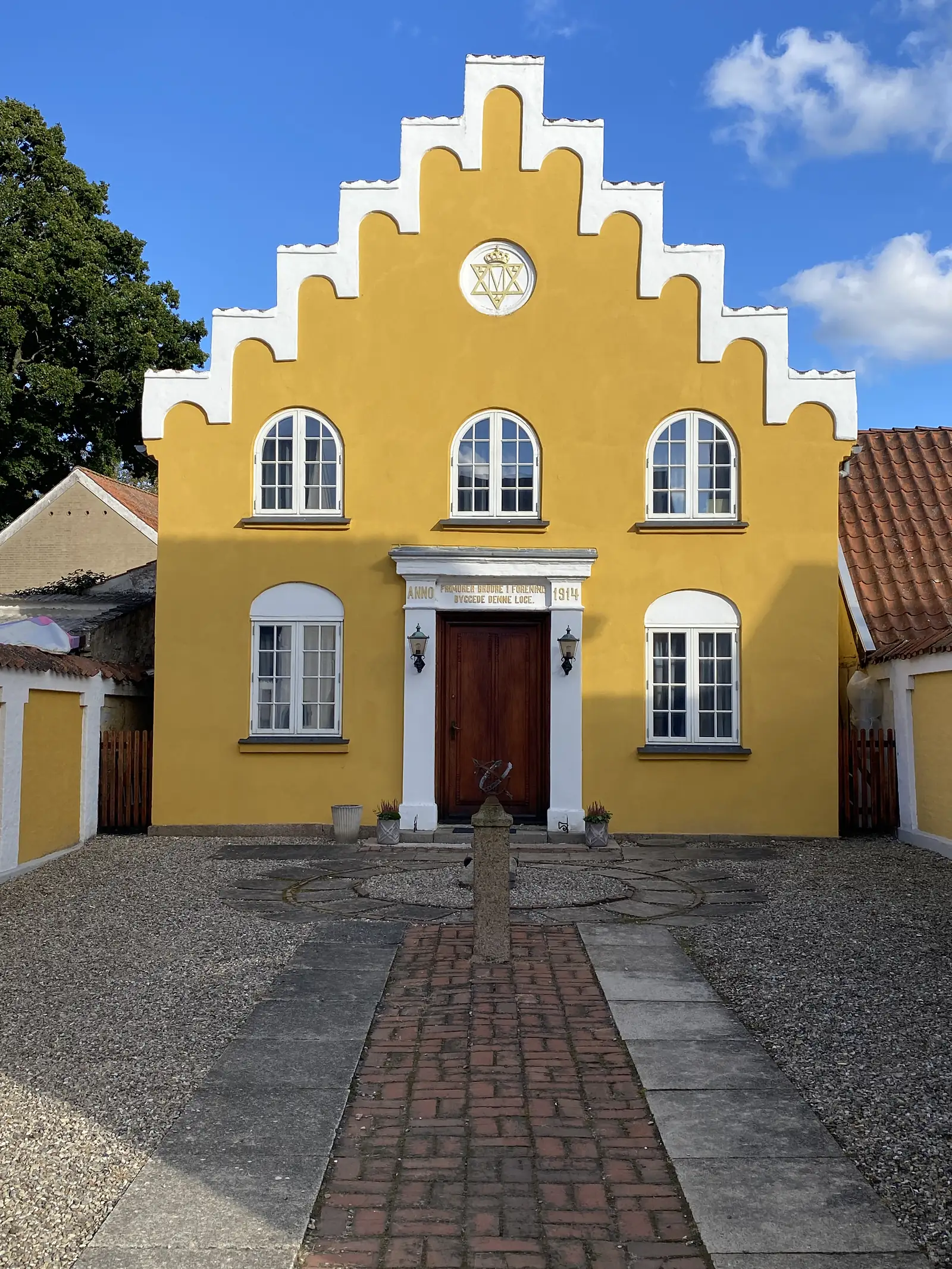 A yellow-rendered building with a white crow-stepped gable and an emblem in a roundel, its doorway dated 1914, at the end of a brick path.