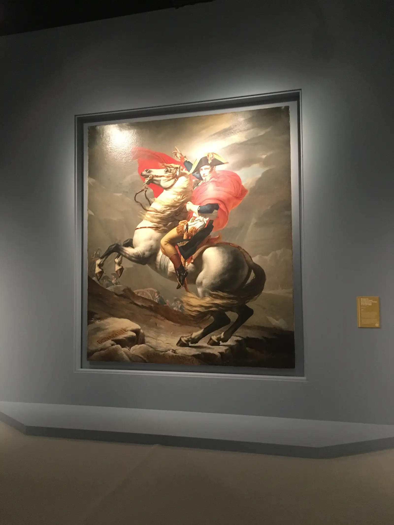 A gallery display of a painted copy of Napoleon crossing the Alps on a rearing white horse, wrapped in a red cloak, with 'BONAPARTE' inscribed on the rock below.