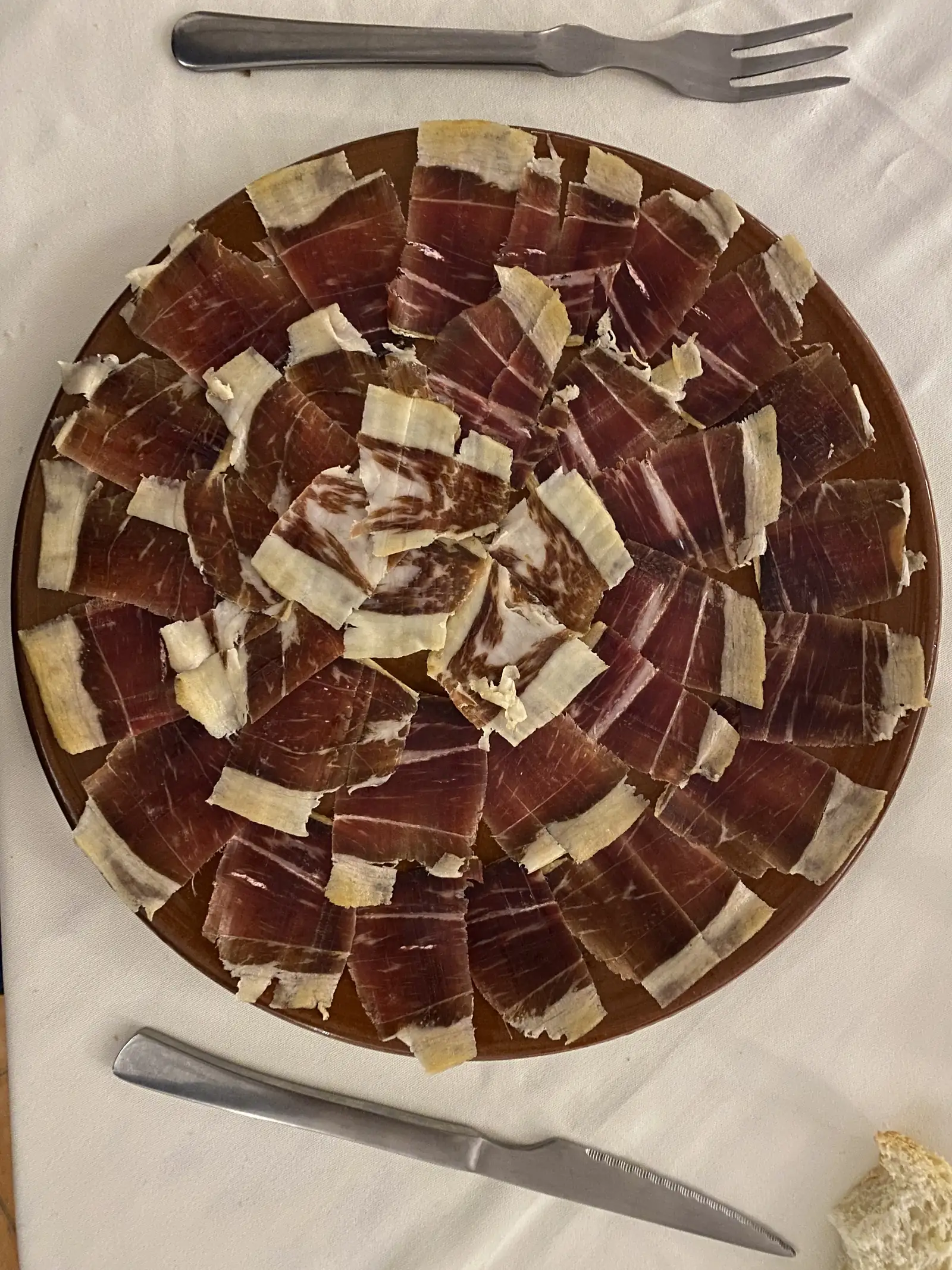 A large terracotta platter spread with thin slices of cured ham on a white tablecloth, with a knife, fork and bread alongside.
