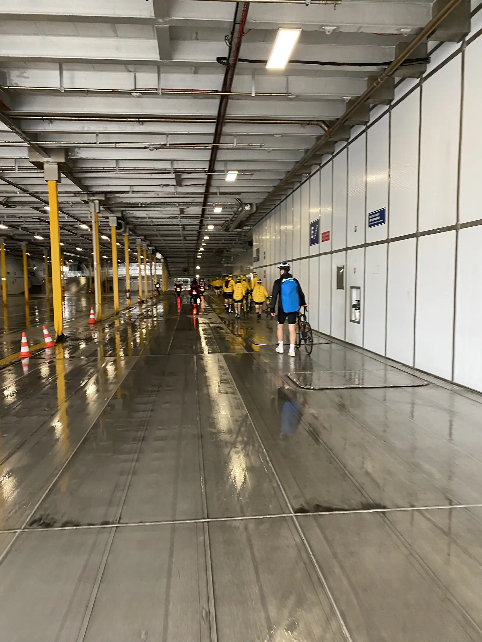 Cyclists wheeling their bikes along the wet vehicle deck of a ferry, a group in yellow jackets ahead under strip lights.