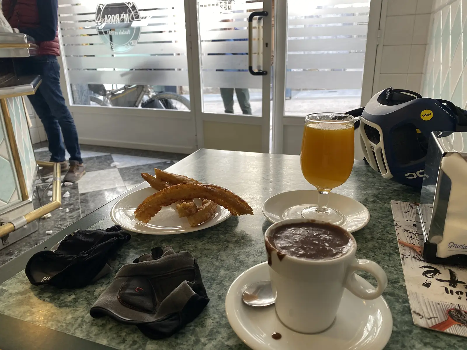 A plate of churros, a glass of orange juice and a cup of thick hot chocolate on a marble cafe table, with cycling gloves and a helmet beside them and the bike outside the window.