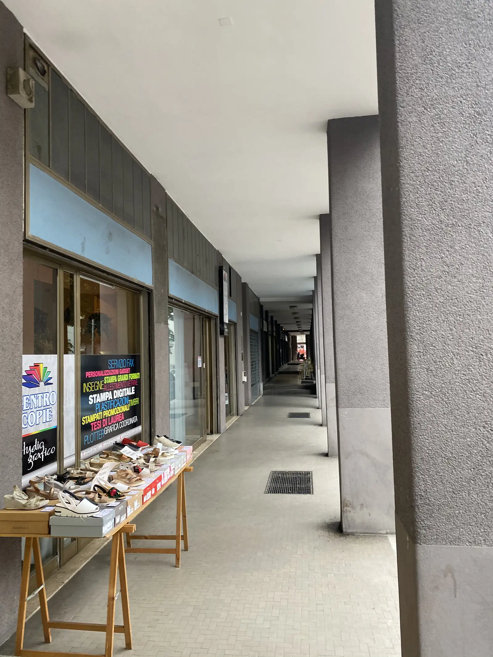 A covered arcade walkway lined with shopfronts, one displaying rows of sandals for sale on trestle tables beneath the colonnade.
