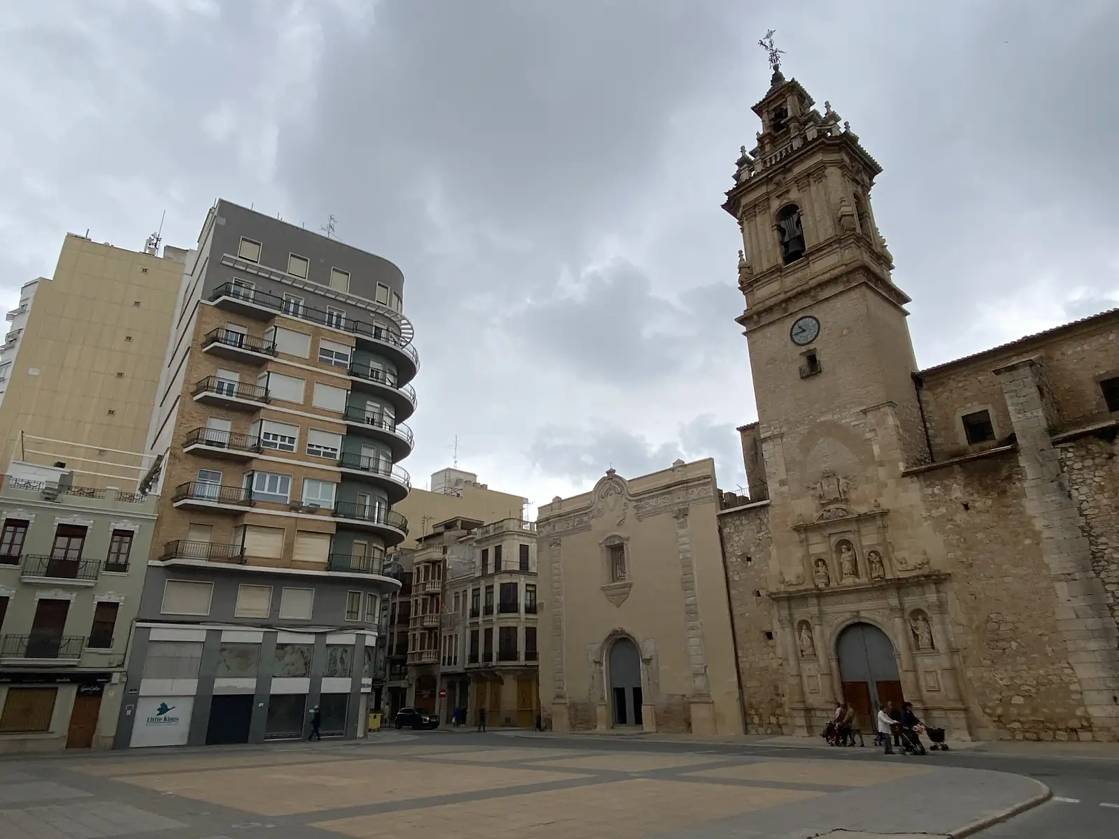 A wide paved town square with a tall baroque bell tower and clock beside a plain chapel front, a curved modern apartment block opposite.