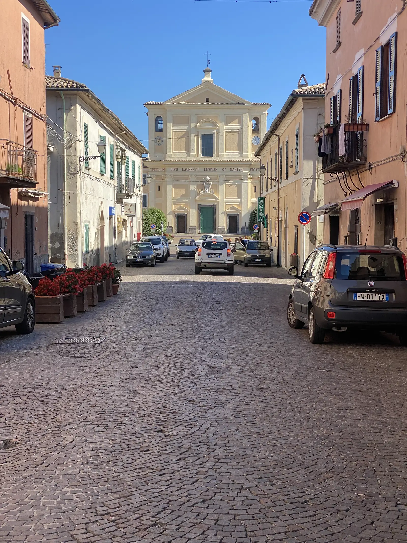 A pale church facade inscribed "TEMPLUM DIVO LAURENTIO LEVITAE ET MARTYRI DICAT..." overlooks a cobbled Italian square lined with parked cars.