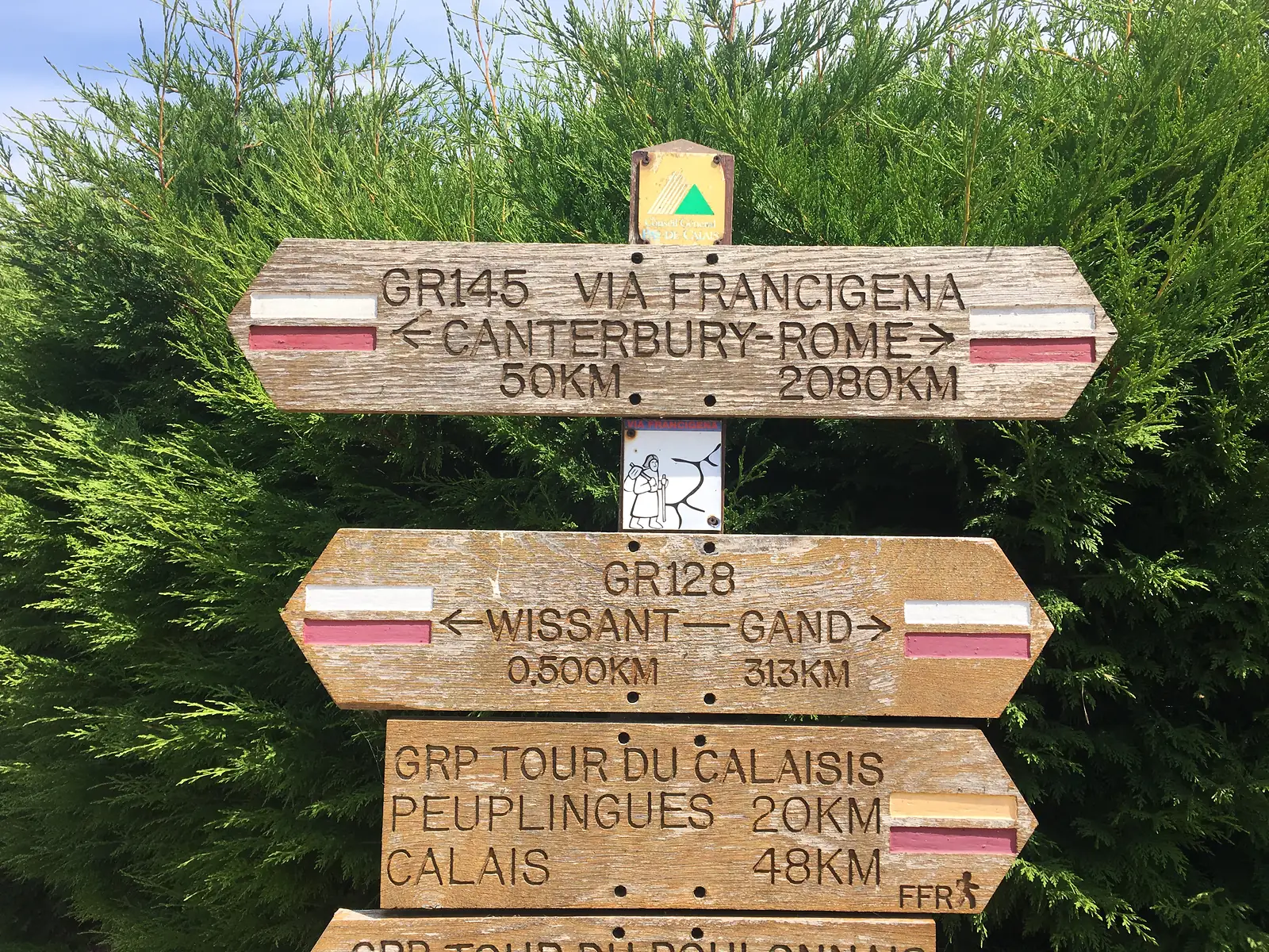 A wooden Via Francigena waymarker reading 'GR145 Via Francigena, Canterbury–Rome, 50km / 2080km', with further signs for GR128 to Wissant and Gand and the GRP Tour du Calaisis to Calais.