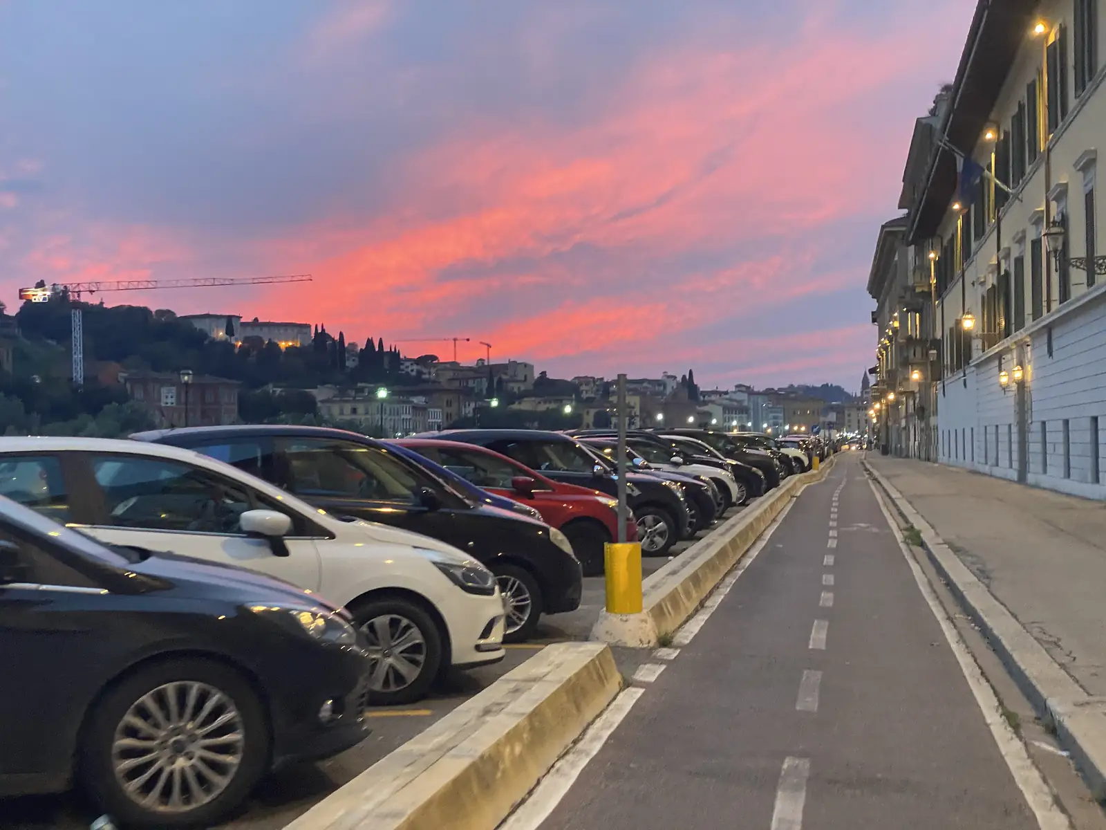 Cars parked along a riverside boulevard beneath a vivid pink and orange sunset over a hillside town with a construction crane.