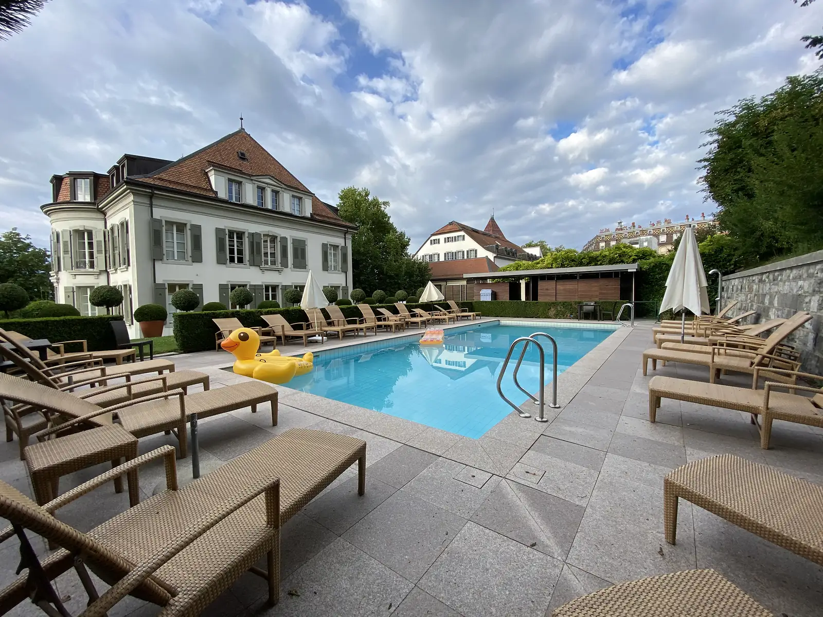 A hotel swimming pool with a giant inflatable yellow duck float, ringed by wicker sun loungers in front of a grand shuttered villa.
