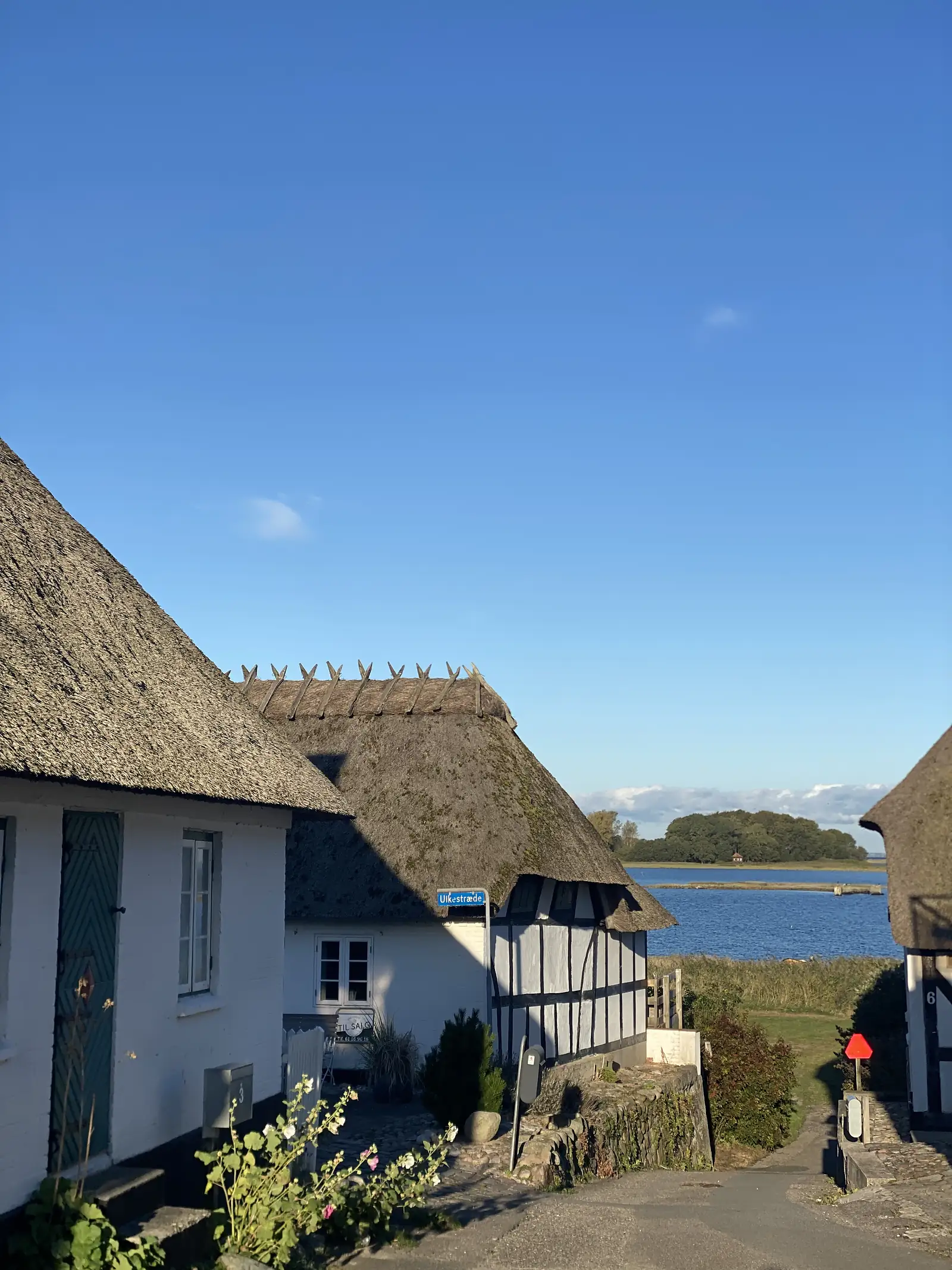 Thatched white and half-timbered cottages at the end of a lane above a bright blue inlet, with a street sign reading "Ulkestræde".