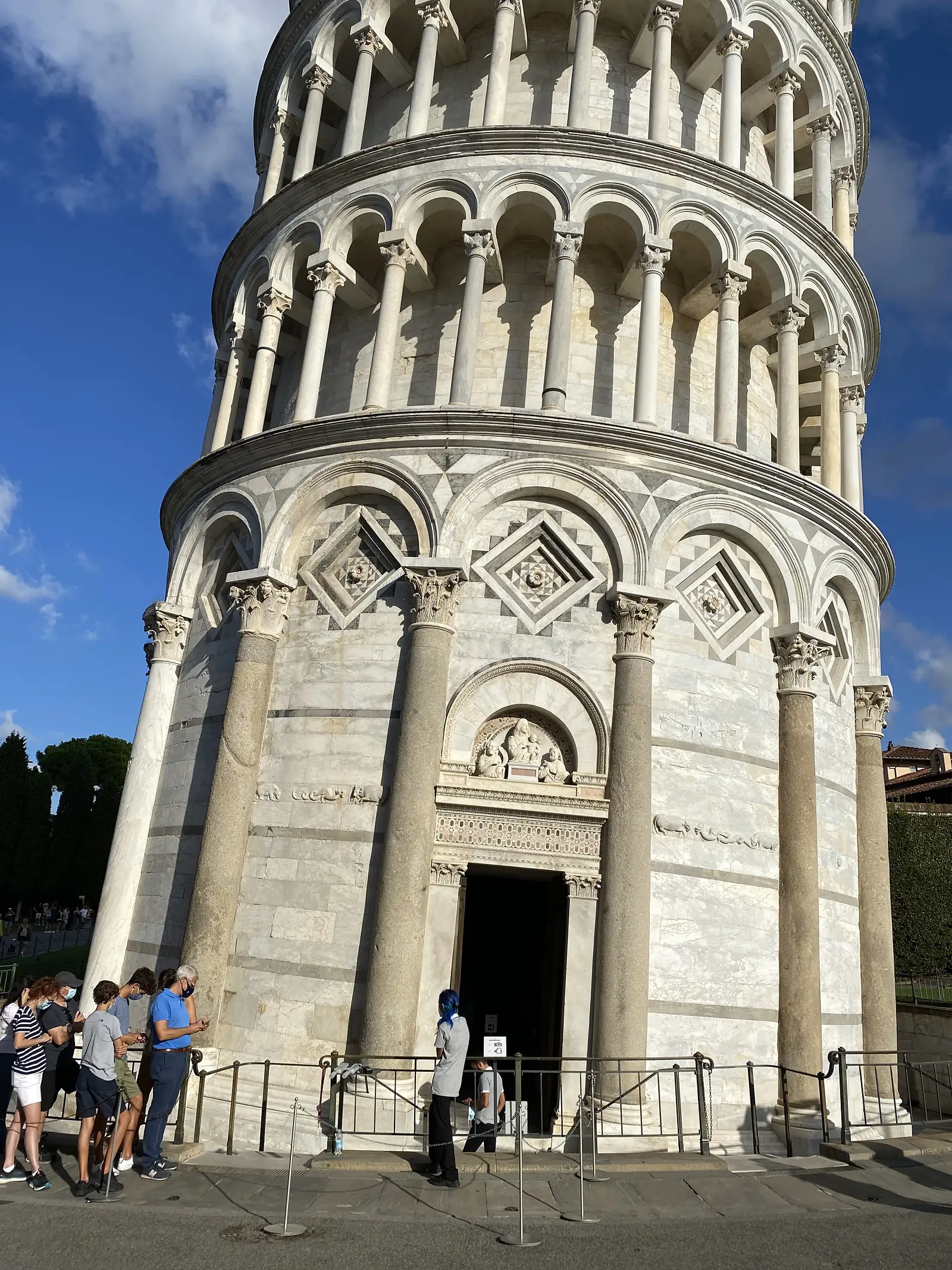 Masked tourists wait in a roped queue at the Baptistery's entrance, its Corinthian columns and diamond marble panels rising above them.
