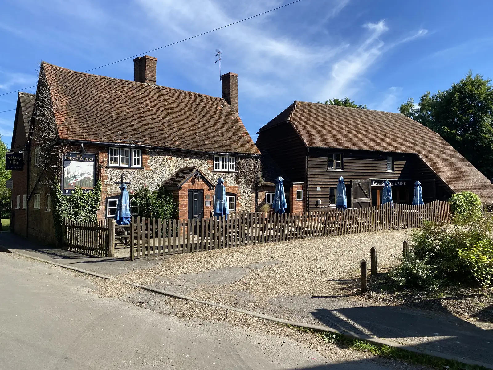 A brick, flint and weatherboarded country pub lettered "THE PERCH & PIKE" with blue furled parasols behind a picket fence.