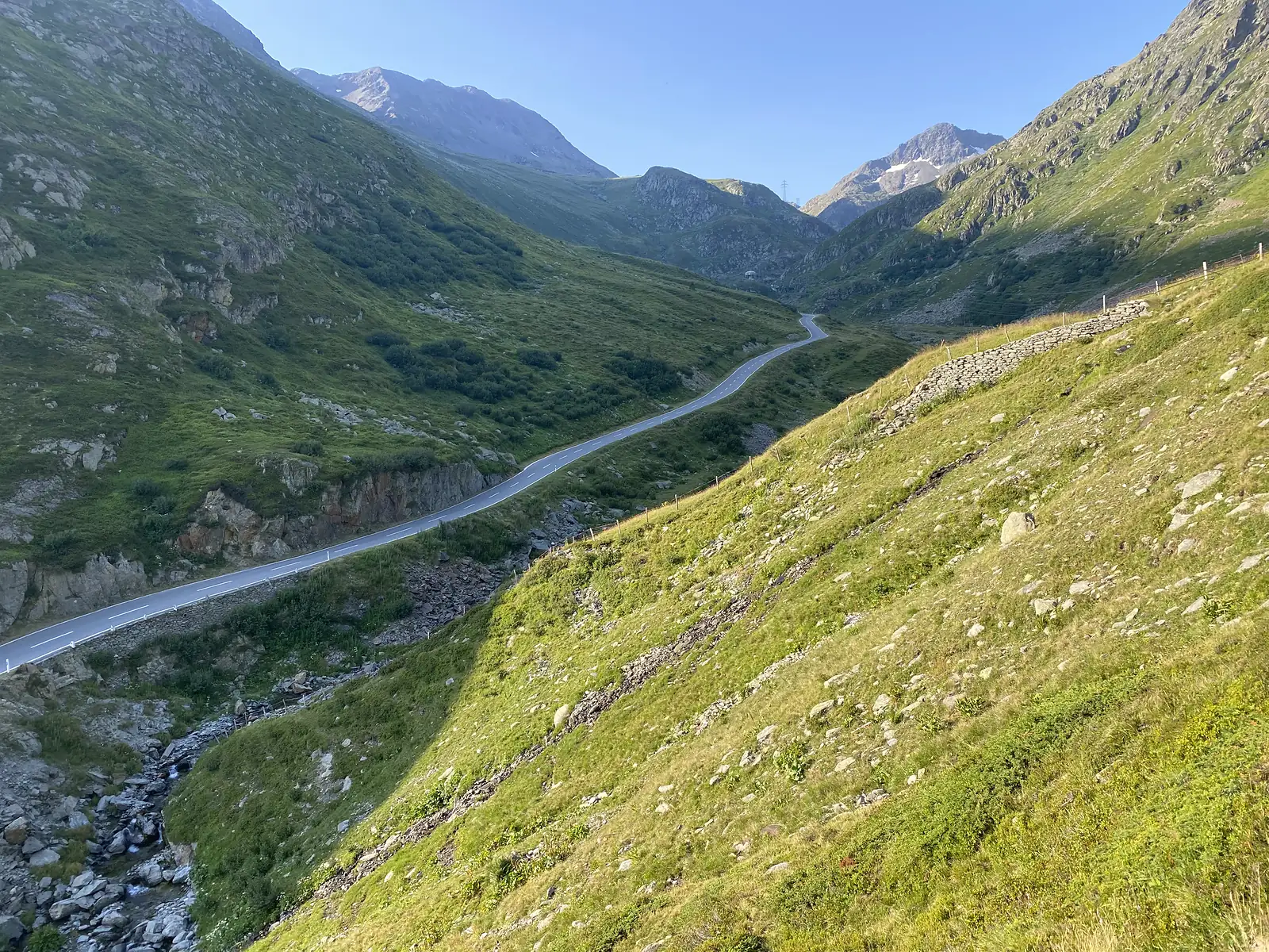 A tarmac road winds down through a green mountain valley towards distant peaks.